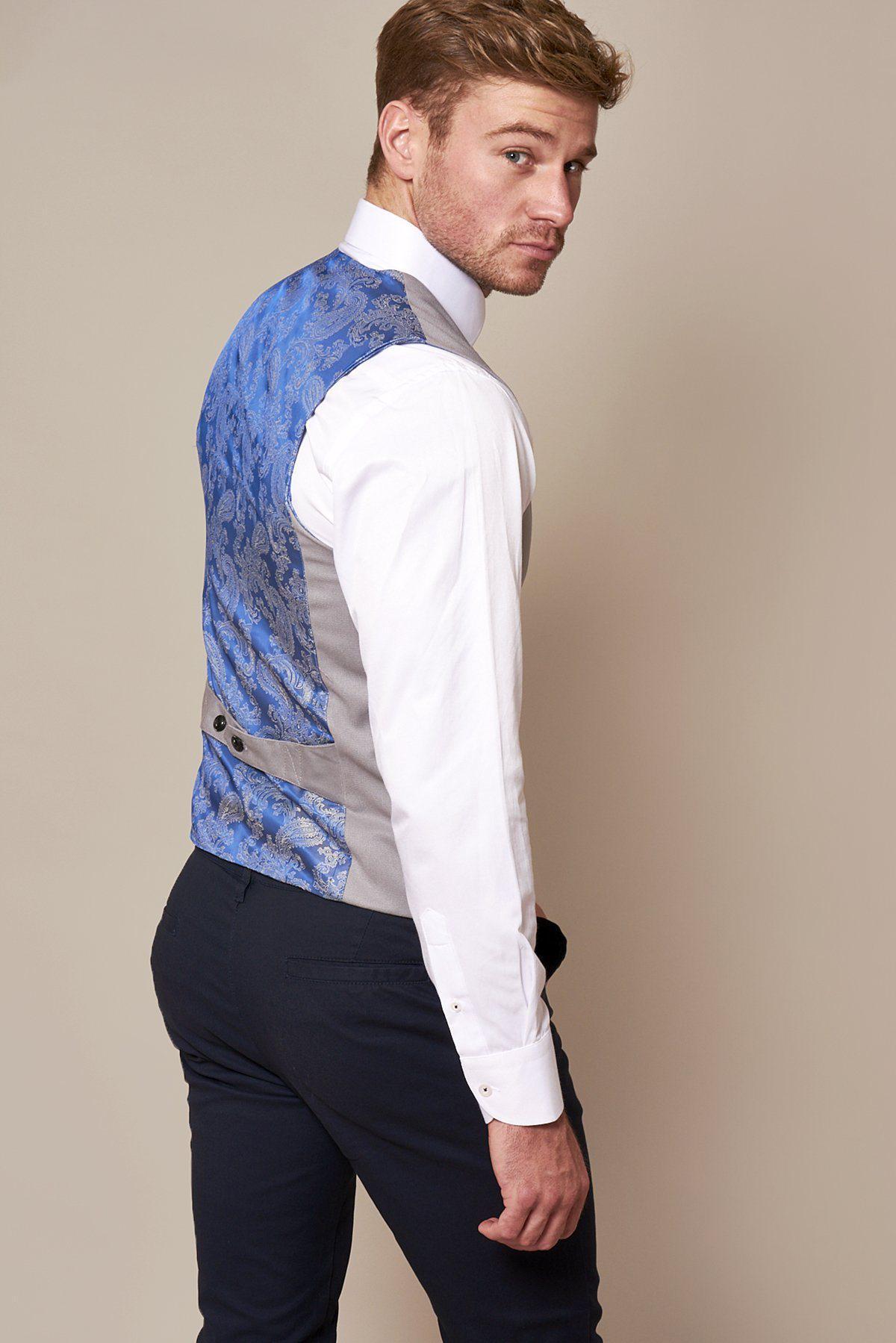 A man with light brown hair wears a marcdarcy KELLY Silver Grey Single Breasted Waistcoat over a white shirt and dark pants, standing turned away from the camera, looking back over his shoulder against a beige background.