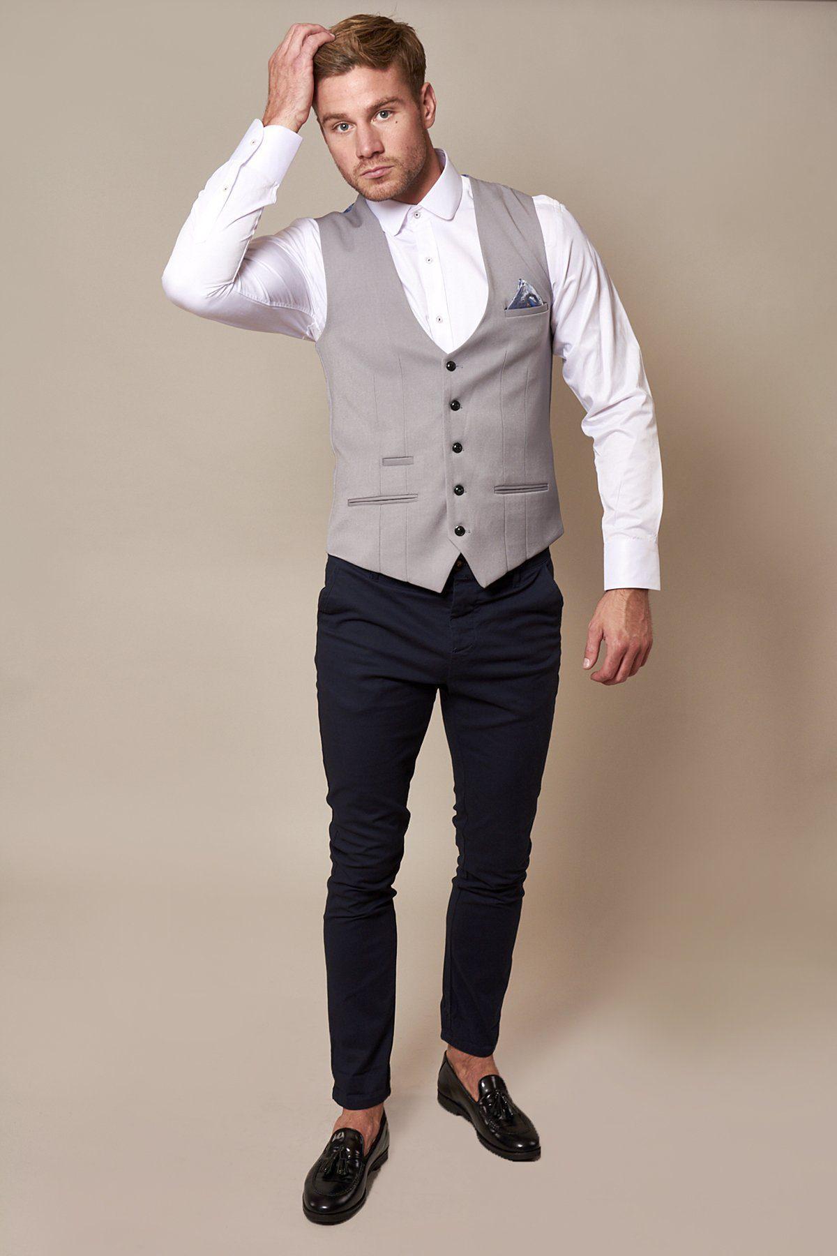 A man with blonde hair wears the marcdarcy KELLY Silver Grey Single Breasted Waistcoat over a white shirt, paired with navy trousers and black loafers, while touching his hair with his hand.