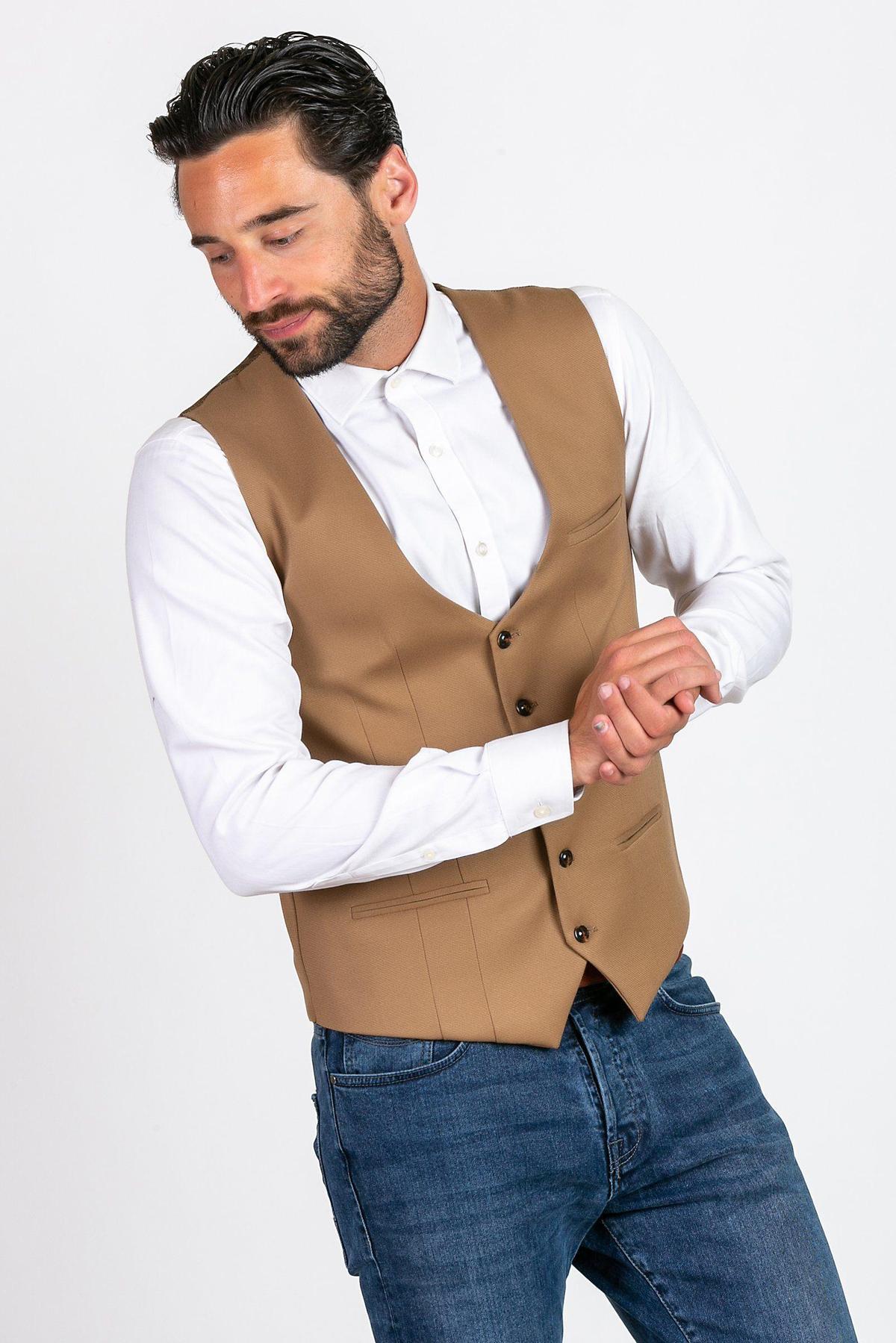 A man with dark hair and a beard wears a white dress shirt, blue jeans, and the marcdarcy KELLY - Tan Single Breasted Waistcoat. He looks down, clasping his hands.