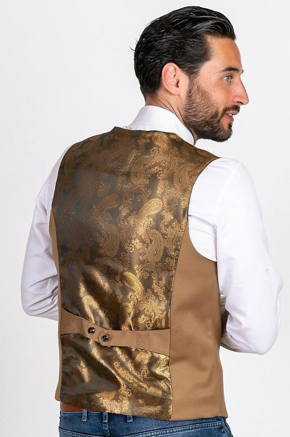 A man with dark hair and a beard wears the marcdarcy KELLY Tan Single Breasted Waistcoat, featuring a gold-brown paisley pattern and adjustable strap, over a white shirt, standing slightly turned against a plain background.
