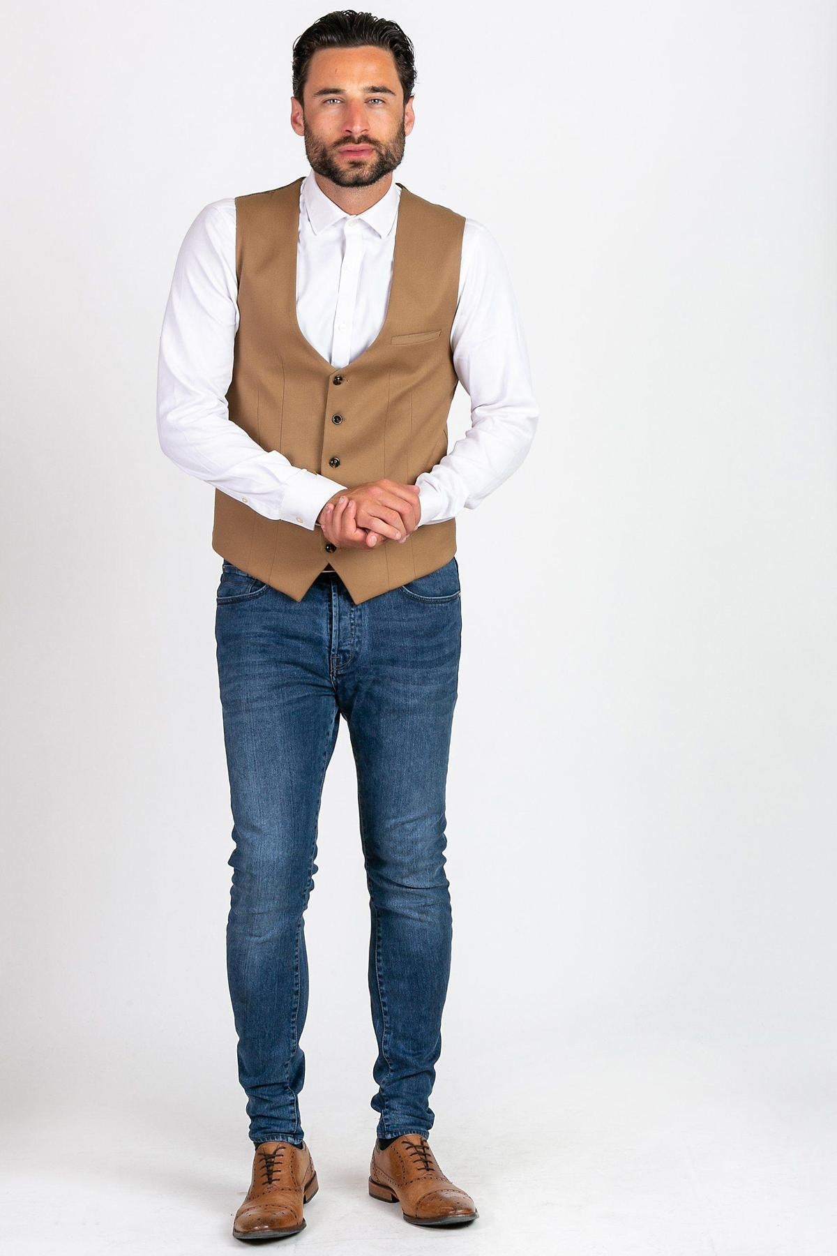 A man stands against a white background wearing the marcdarcy KELLY Tan Single Breasted Waistcoat over a white shirt, paired with blue jeans and brown dress shoes, his hands clasped in front of him.