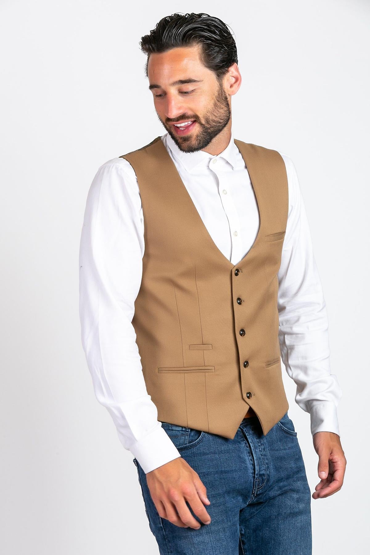 A man with short dark hair and a beard wears the marcdarcy KELLY Tan Single Breasted Waistcoat over a white dress shirt and blue jeans, looking down and slightly to the side against a plain white background.