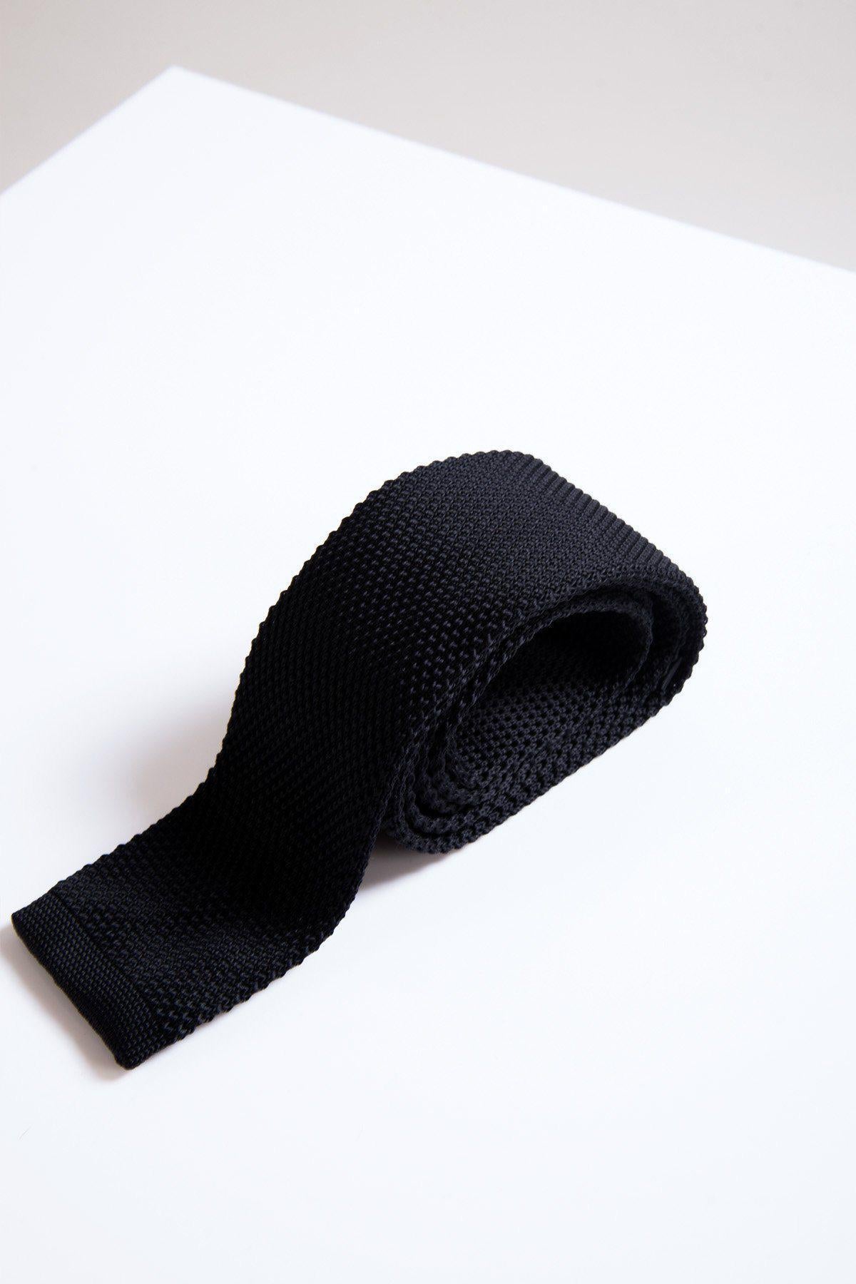 The KT - Black Knitted Tie by Accessory, a stylish formal wear item, is rolled up and displayed on a white surface against a light gray background.