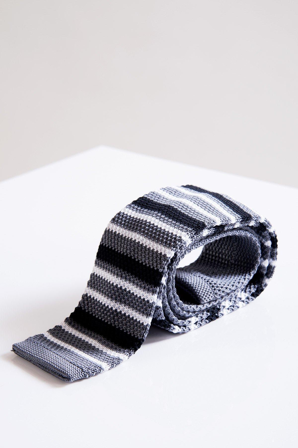 The Accessory KT - Grey Stripe Knitted Tie, featuring horizontal black, white, and grey stripes, is displayed rolled up on a white surface to highlight its modern knit design and lightweight fabric.