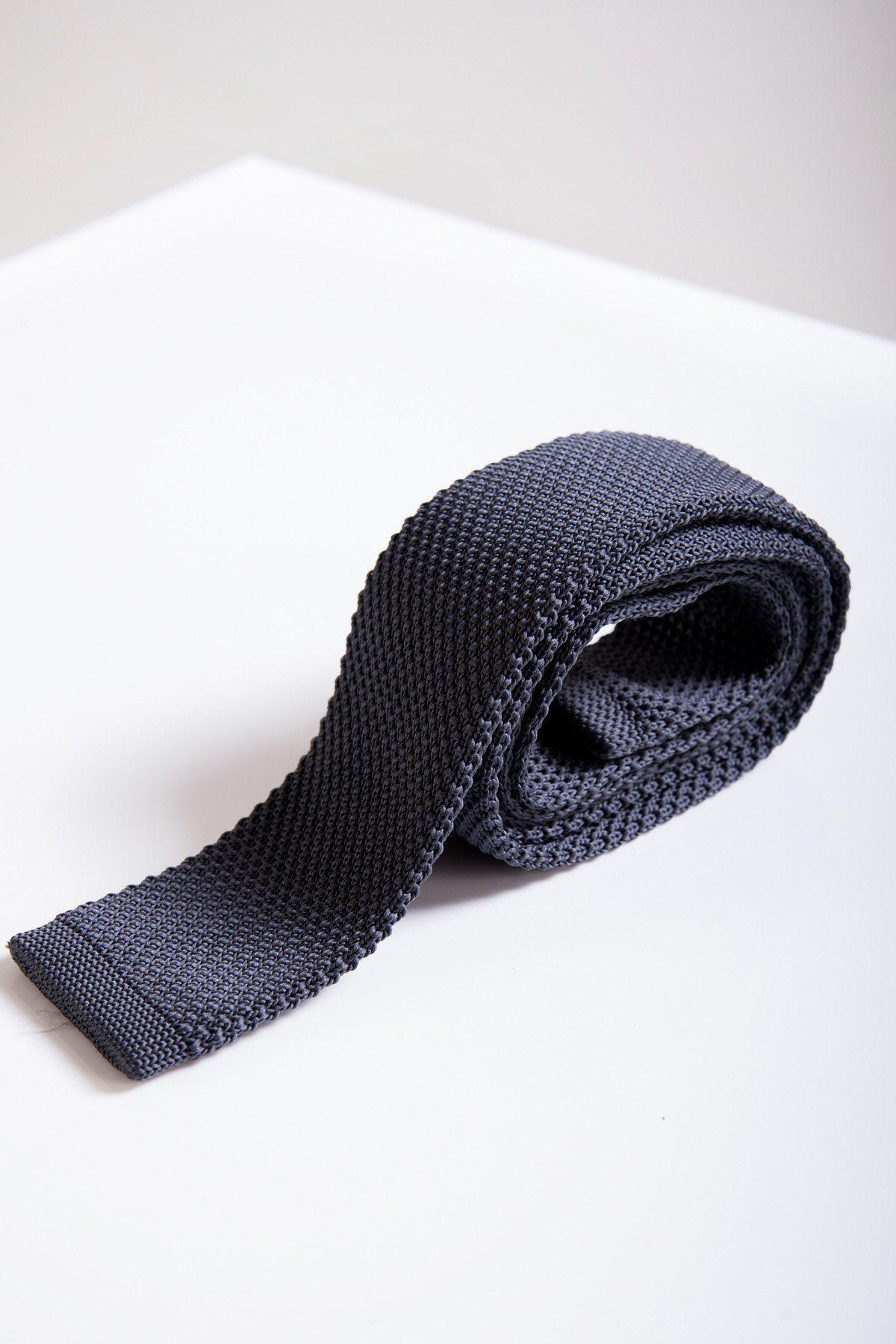 The Accessory KT - Mid Grey Knitted Tie, featuring a modern knit design and squared end, is rolled up on a white surface—its textured fabric making it ideal for pairing with your office suit.