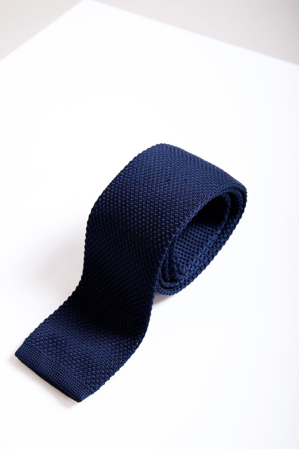 The KT - Navy Knitted Tie by Accessory, featuring a textured design, is rolled up and placed on a white surface with a soft shadow, its tip pointing toward the bottom left corner.