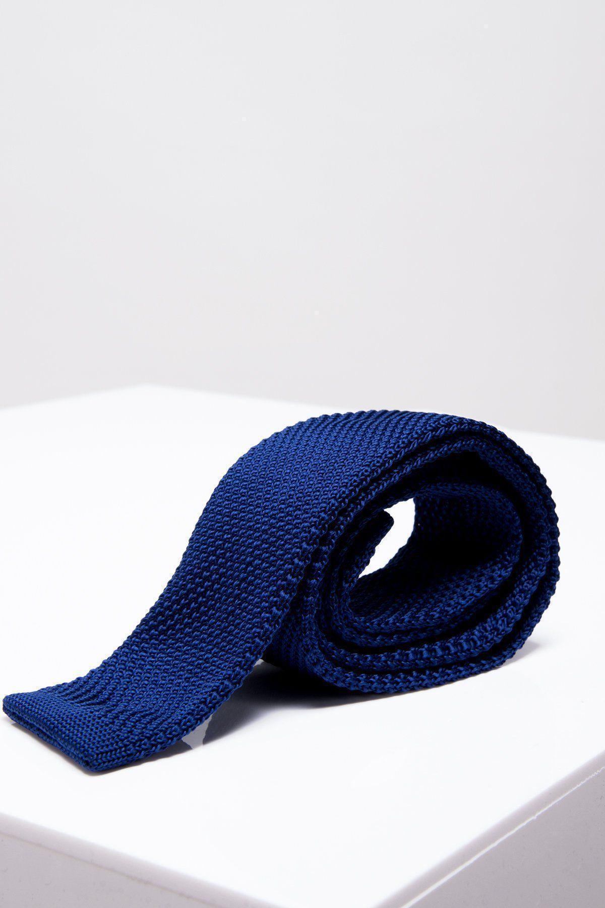 The Accessory KT - Royal Blue Knitted Tie, featuring a modern textured design, is rolled up and displayed on a glossy white surface against a plain light background.