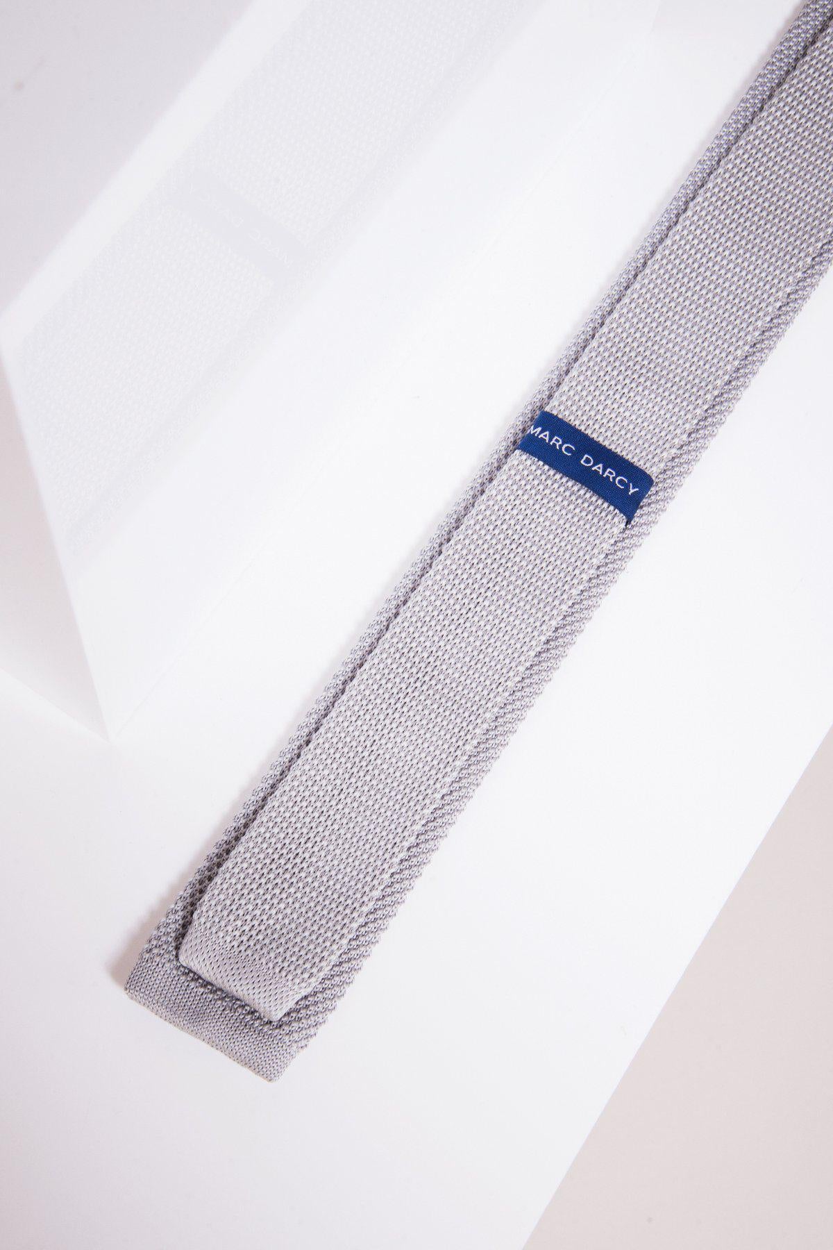 The KT - Silver Grey Knitted Tie by Accessory features a modern textured design and blue label, shown on a white surface with its lightweight fabric catching geometric shadows.