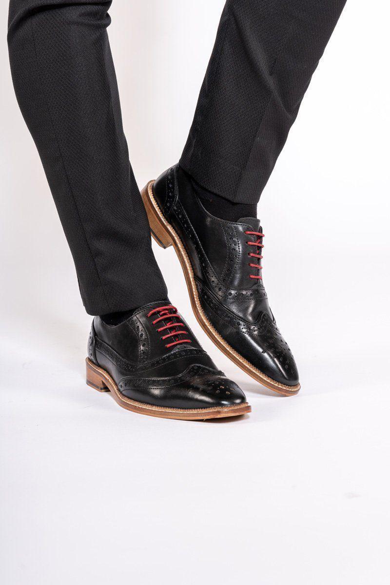 A person stands against a white background, wearing black dress pants and Marc Darcys LARKIN Black Leather Brogue Shoes featuring red laces, tan soles, and a hand-crafted design.