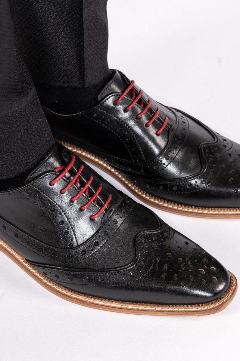 A person wearing Marc Darcys LARKIN Black Leather Brogue Shoes with red laces and light brown soles, paired with black dress pants, stands on a white surface.