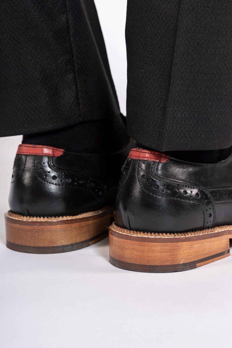 Close-up of a person wearing Marc Darcy’s LARKIN Black Leather Brogue Shoes, featuring brown soles and red heel accents. The handcrafted shoes are paired with black pants and positioned on a white surface.
