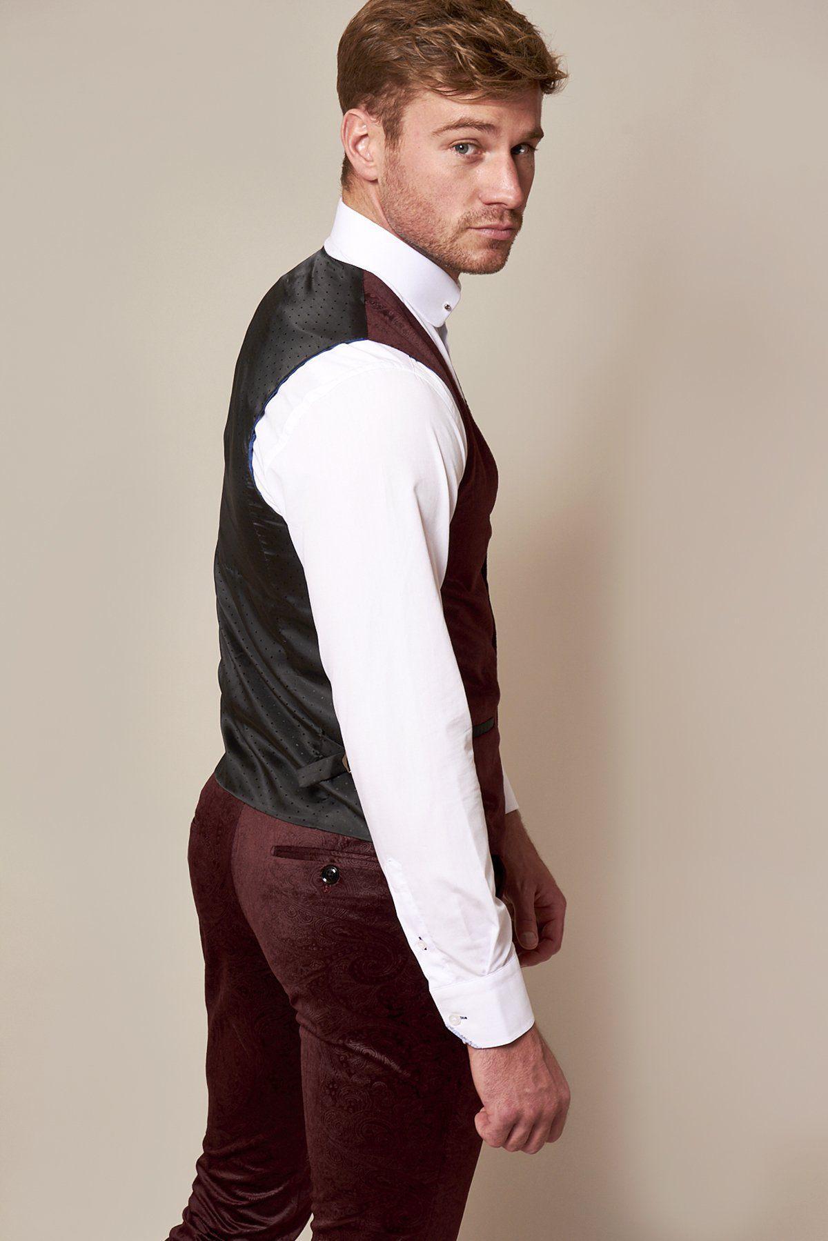 A man with short light brown hair poses in a slim fit Simon Wine Velvet Suit by Marc Darcy, inspired by Fenerbahçe and Brazil midfielder Fred, pairing it with a black vest and white shirt against a plain beige background.