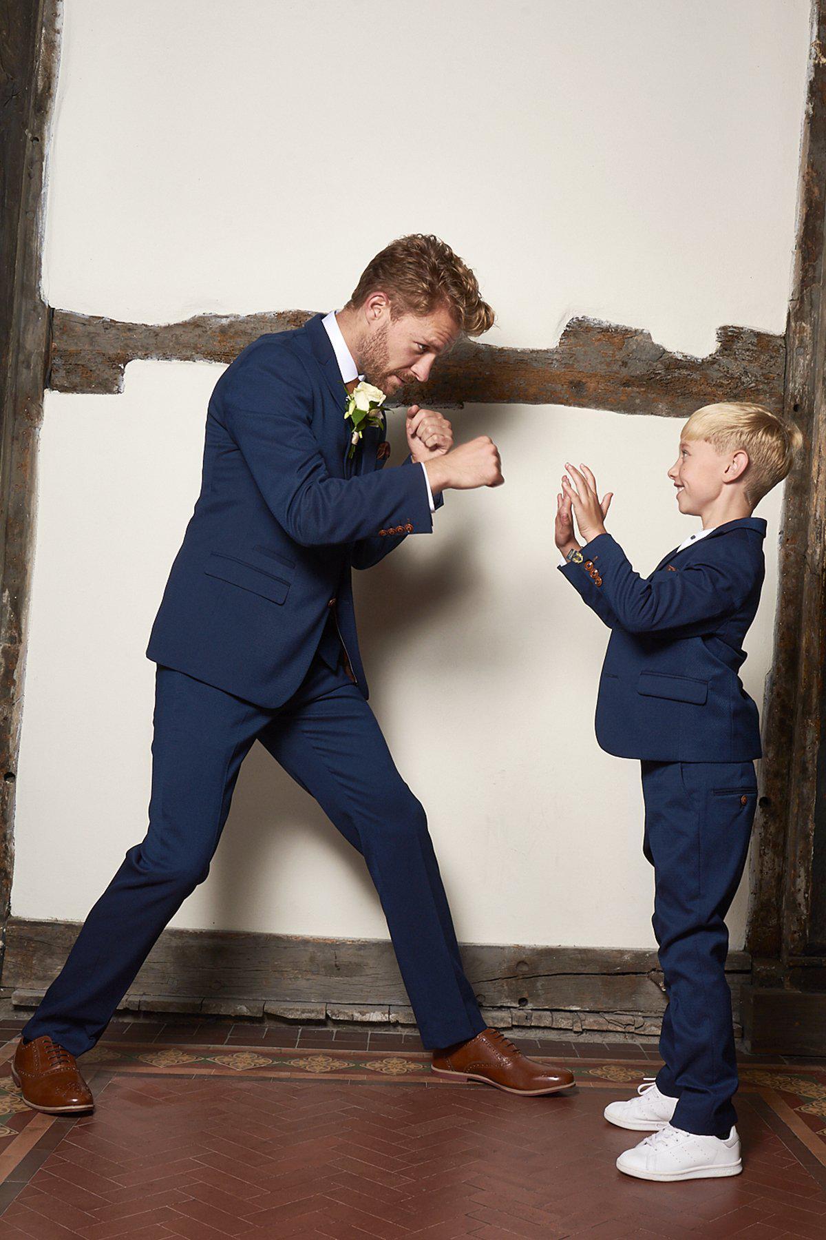 A man and a boy, both wearing the marcdarcy Matching Father & Son | Men’s MAX Royal Blue Three Piece Suit, smile and playfully spar together in a room with wooden beams and patterned tile flooring.