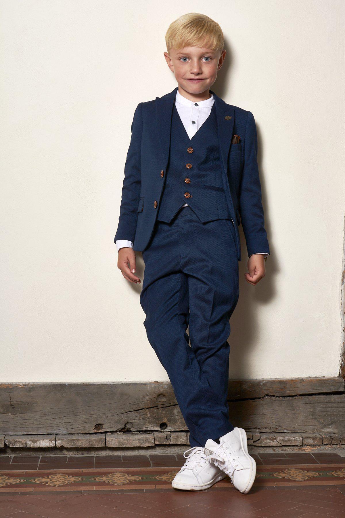 A young boy in a marcdarcy Matching Father & Son | Men’s MAX Royal Blue Three Piece Suit stands casually against a cream wall, wearing the slim-fit blazer over a white shirt and pairing it with white sneakers.