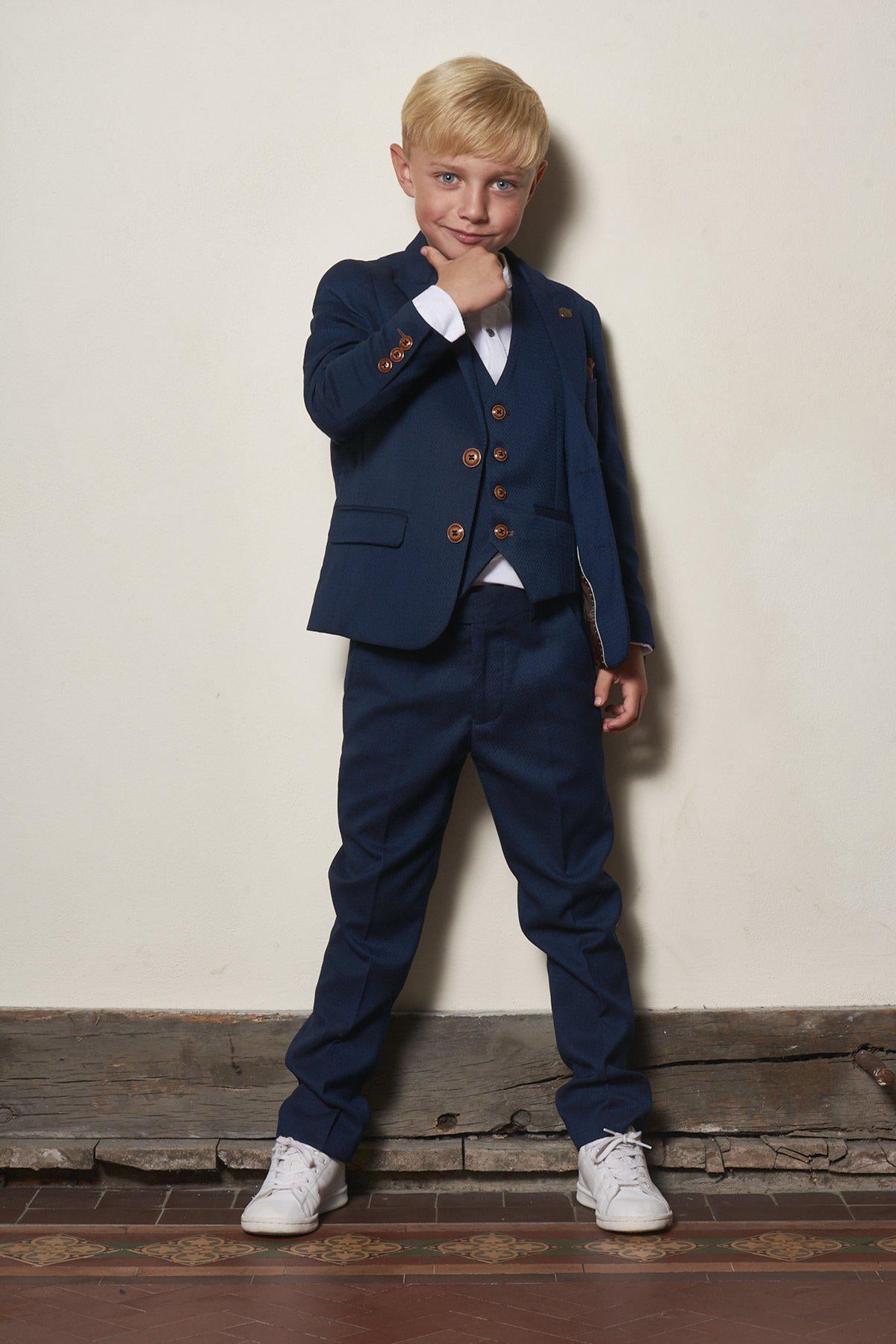 A young boy with blond hair and blue eyes smiles while touching his chin, wearing the marcdarcy Matching Father & Son Men’s MAX Royal Blue Three Piece Suit with a white shirt and white sneakers.