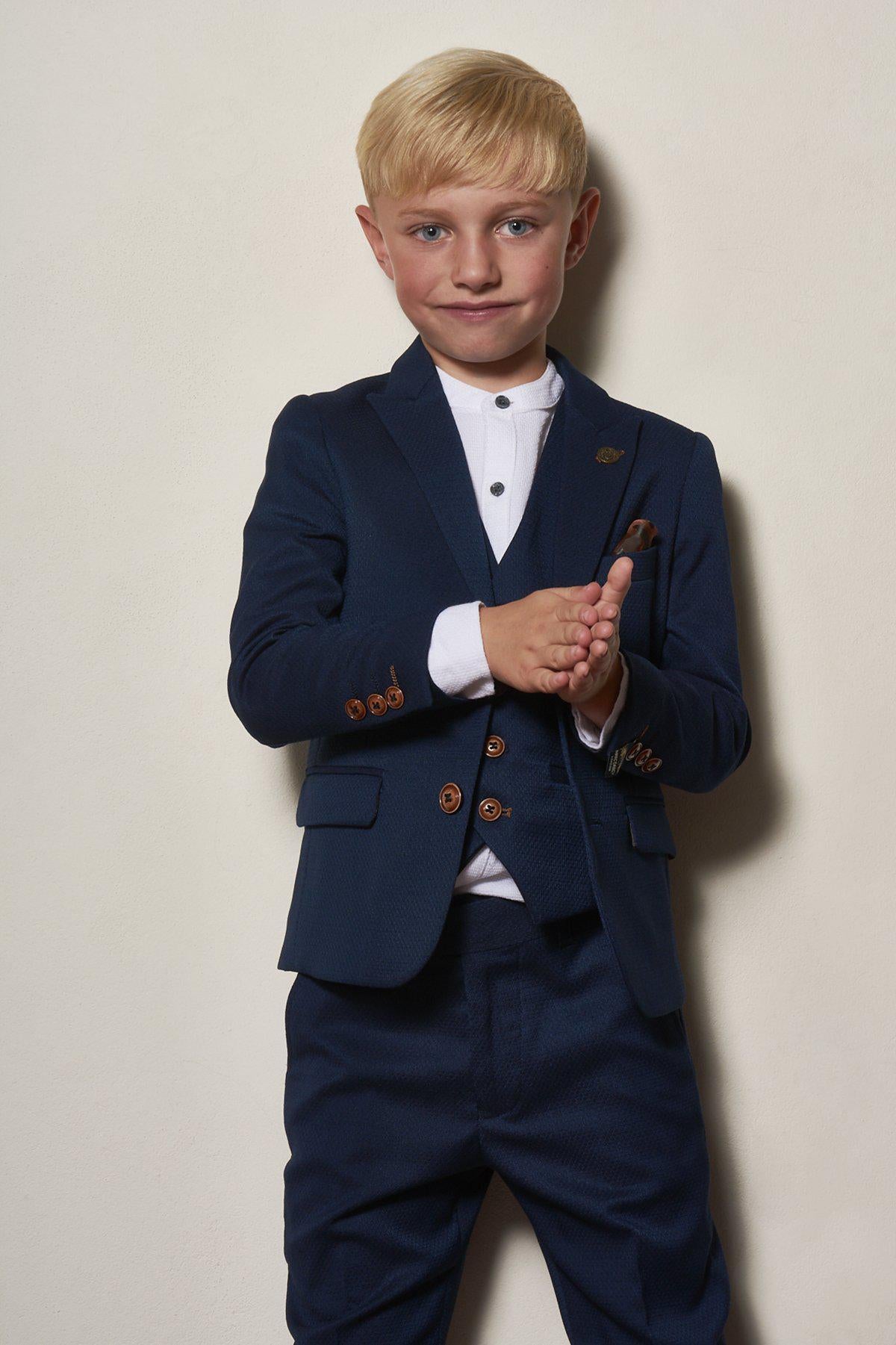 A young boy smiles softly, standing against a plain wall in the marcdarcy Matching Father & Son | Men’s MAX Royal Blue Three Piece Suit, styled with a white shirt and camouflage print pocket square, hands together in front of him.