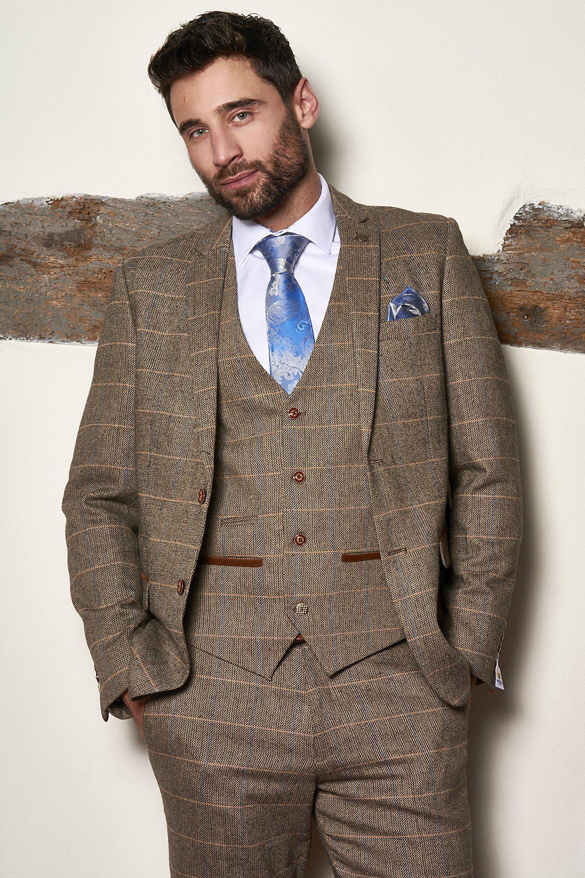 A man with dark hair and a beard leans against a wall, wearing the marcdarcy Matching Father & Son Men’s TED Tan Tweed Check Three Piece Suit, styled with a white shirt and blue patterned tie with matching pocket square.