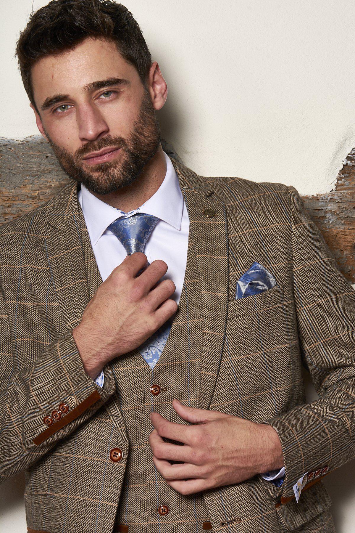 A man with short dark hair and a beard adjusts his tie while wearing the marcdarcy Matching Father & Son Men’s TED Tan Tweed Check Three Piece Suit, standing against a textured wall.