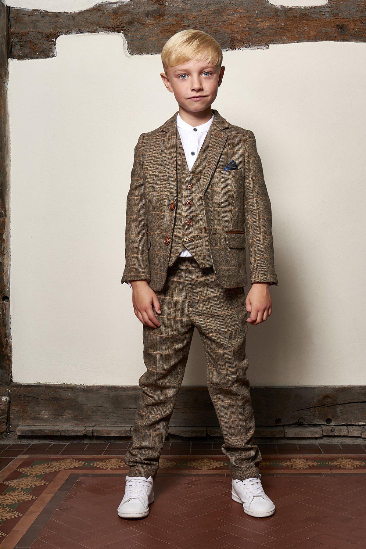 A young boy stands indoors wearing the marcdarcy Matching Father & Son Men’s TED Tan Tweed Check Three Piece Suit, white shirt, and sneakers on a patterned tile floor, posing against a rustic wall and looking at the camera.