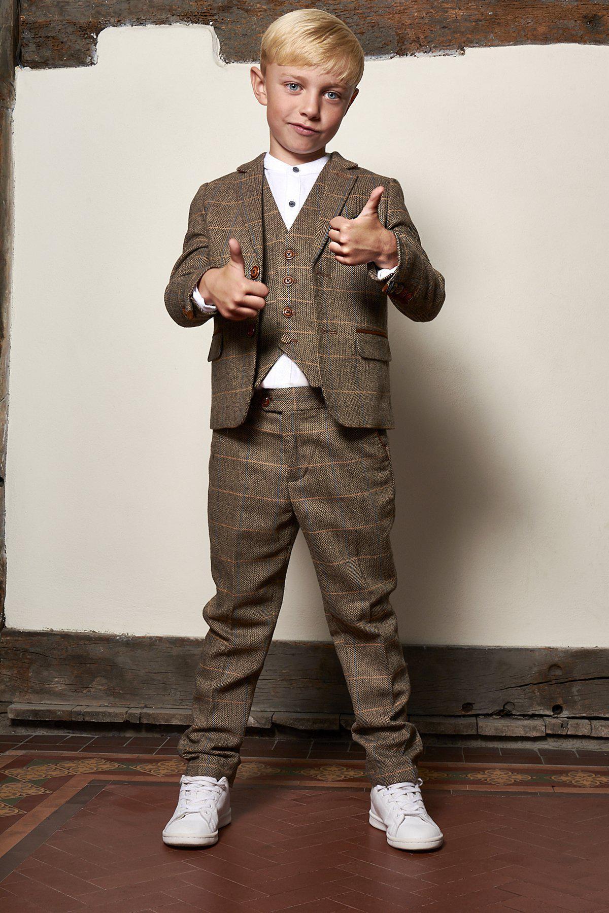 A young boy with blonde hair smiles and gives two thumbs up while wearing the marcdarcy Matching Father & Son Men’s TED Tan Tweed Check Three Piece Suit, paired with white sneakers. He stands indoors against a white wall with wooden trim.