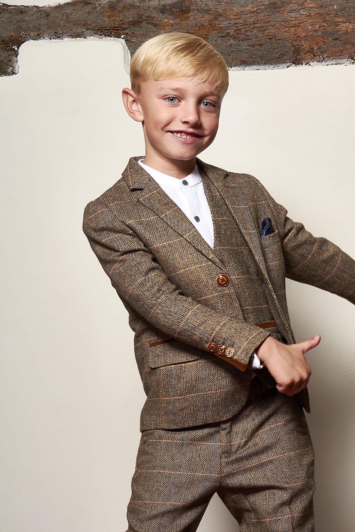 A young boy smiles at the camera, wearing the marcdarcy Matching Father & Son | Men’s TED Tan Tweed Check Three Piece Suit, standing against a cream wall with exposed brick at the top.