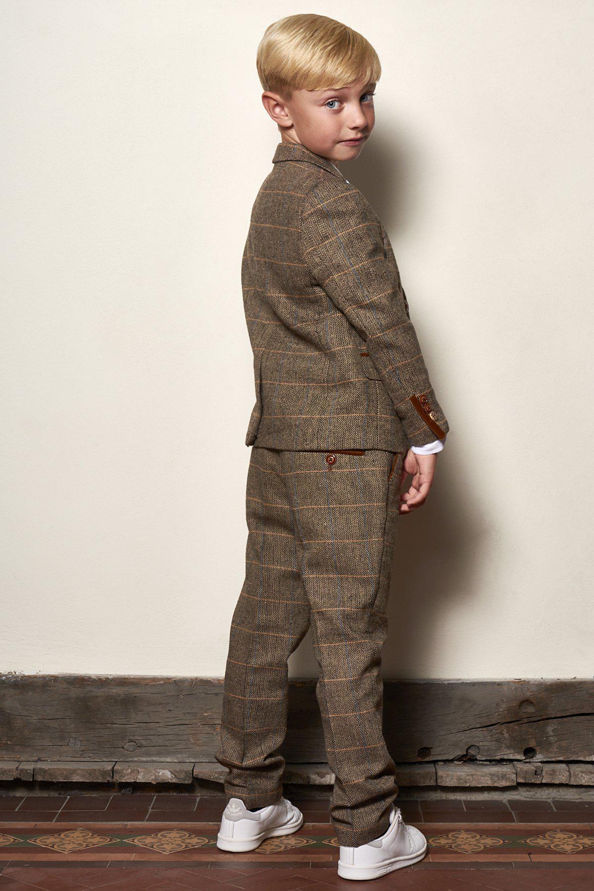 A young blond boy stands indoors on patterned tiles, wearing the marcdarcy Matching Father & Son | Men’s TED Tan Tweed Check Three Piece Suit with white sneakers, glancing over his shoulder at the camera.