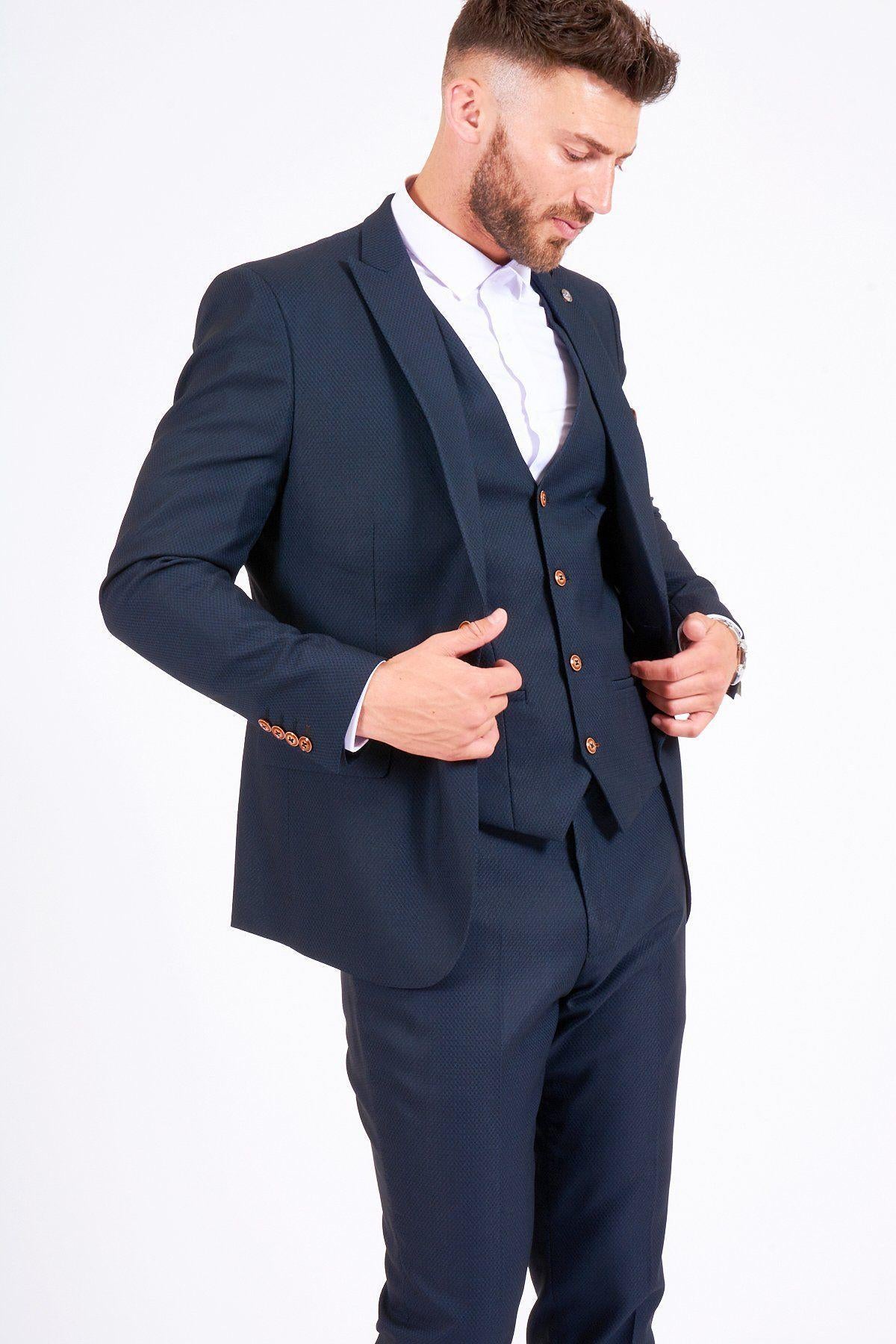 A man wearing the marcdarcy MAX Navy Suit with TED Waistcoat and a white shirt adjusts his jacket while looking down, set against a plain white background.