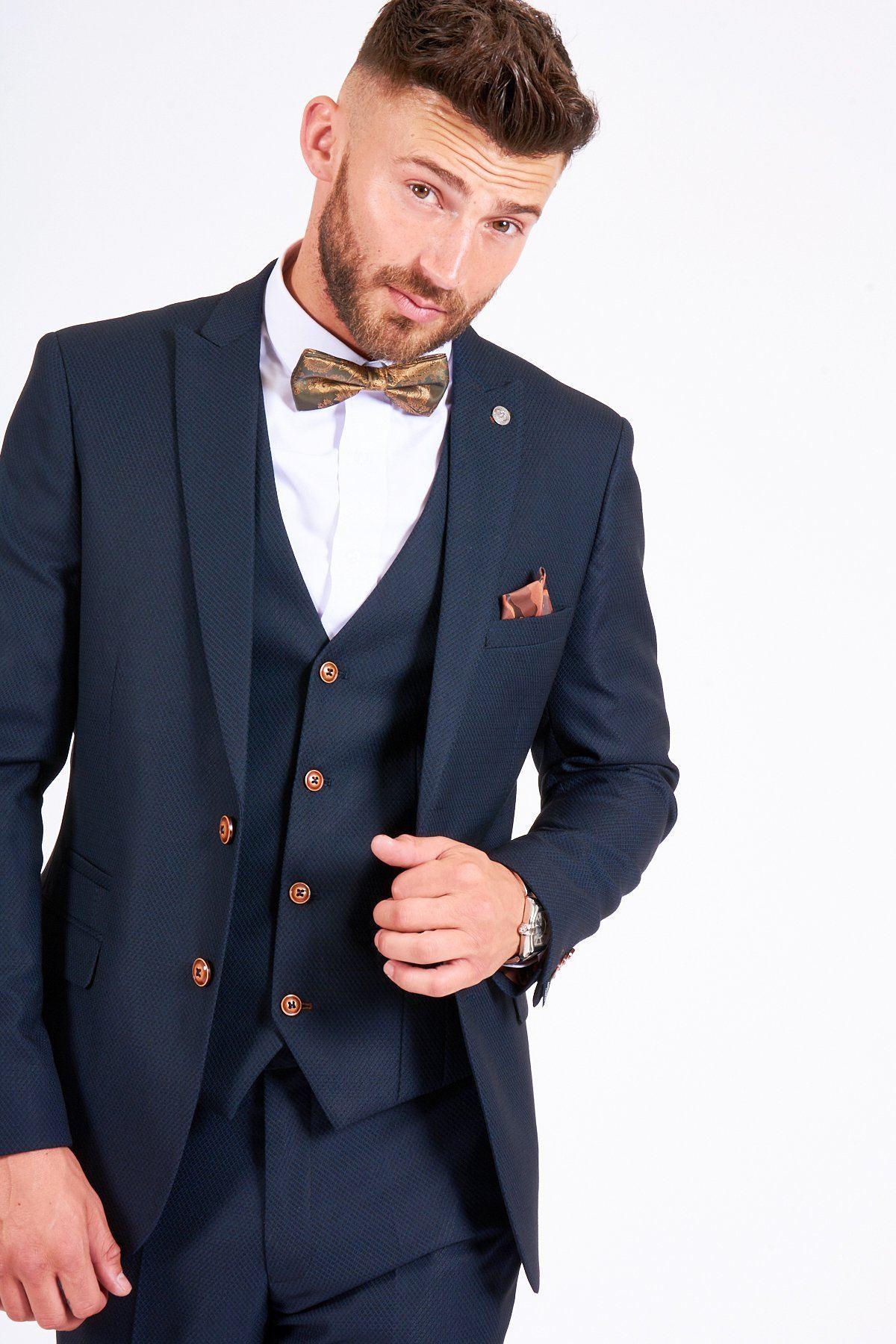A man with short brown hair and a beard wears the marcdarcy MAX navy suit with TED waistcoat, paired with a white shirt, patterned bow tie, and pocket square. He looks at the camera, slightly tilting his head while adjusting his jacket.