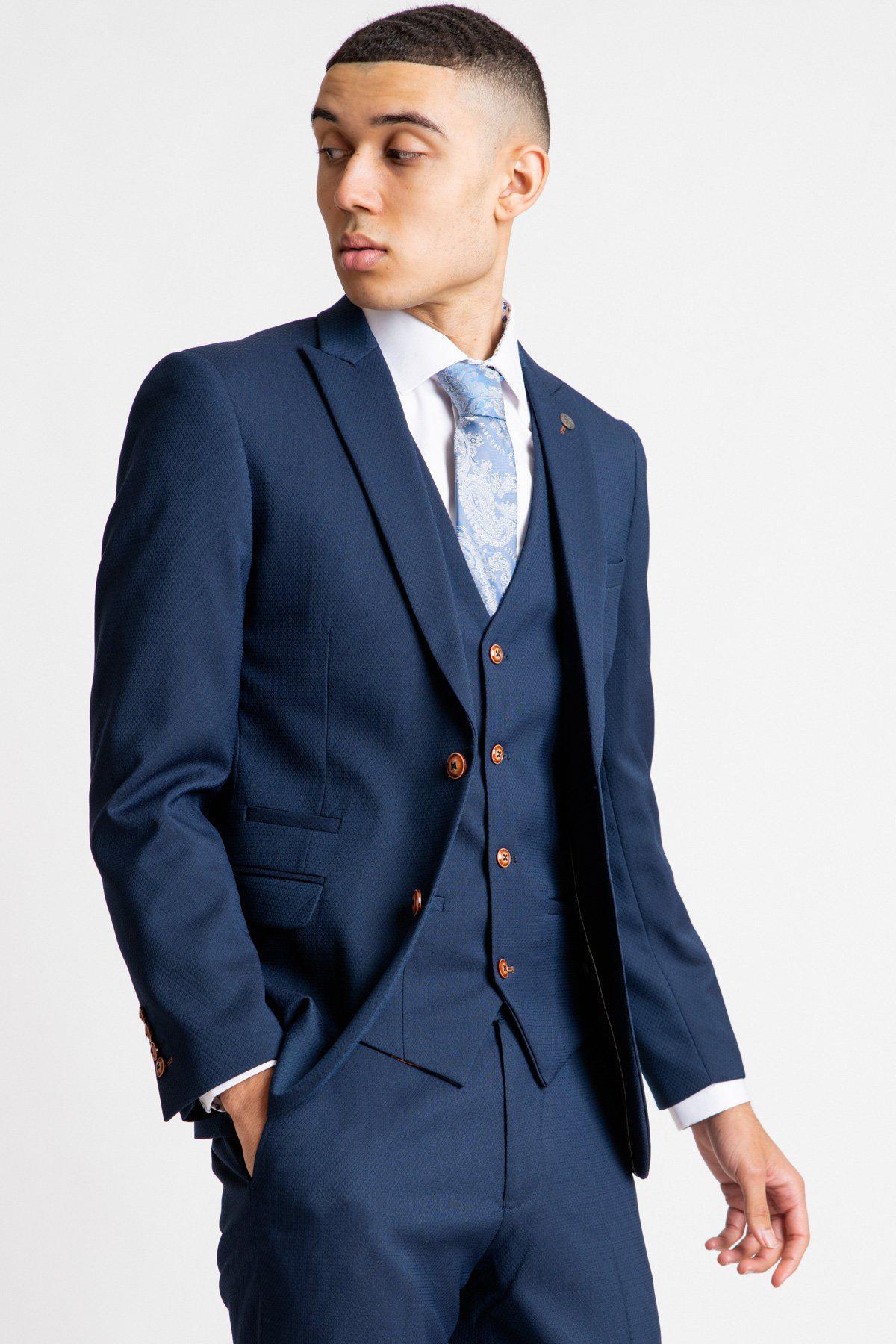 A young man wears the marcdarcy MAX - Royal Blue Blazer with TED Waistcoat, paired with a white shirt and patterned blue tie, looking left with one hand in his pocket against a plain light background.
