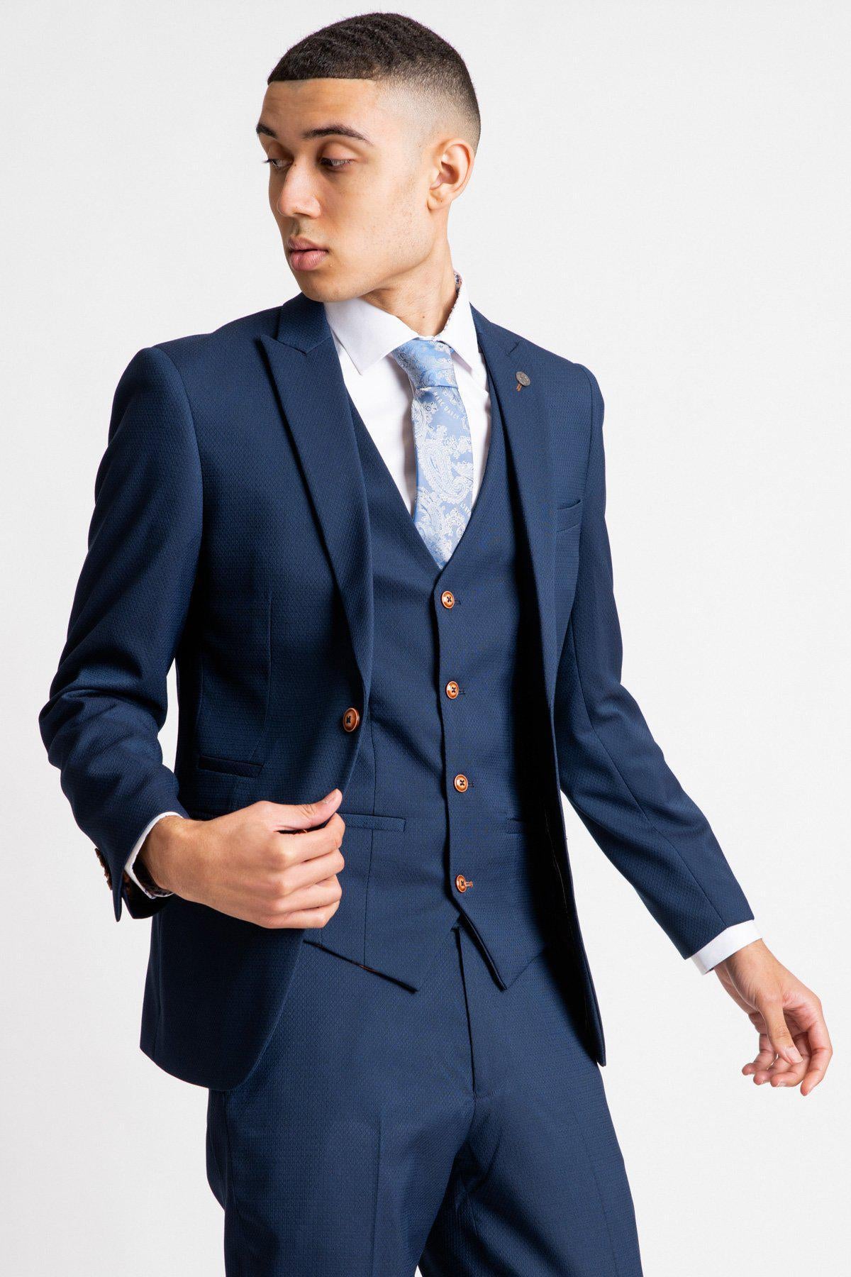 A young man in the marcdarcy MAX - Royal Blue Blazer with TED Waistcoat stands against a white background, looking left while adjusting his double-breasted waistcoat and wearing a light blue floral tie.