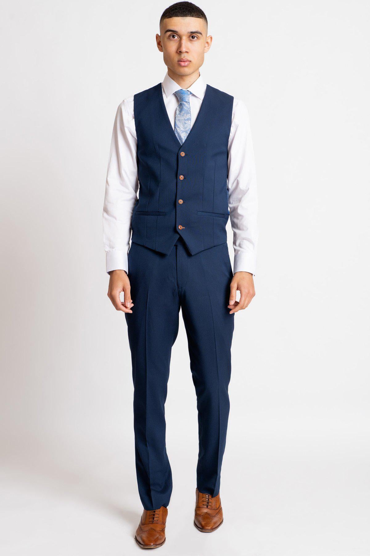 A man wears marcdarcy’s The Everton Collection | Dominic Calvert-Lewin In The MAX Royal Blue Suit, featuring a three-piece royal blue suit with copper buttons, styled with a white shirt, light blue tie, and tan leather Oxford shoes.