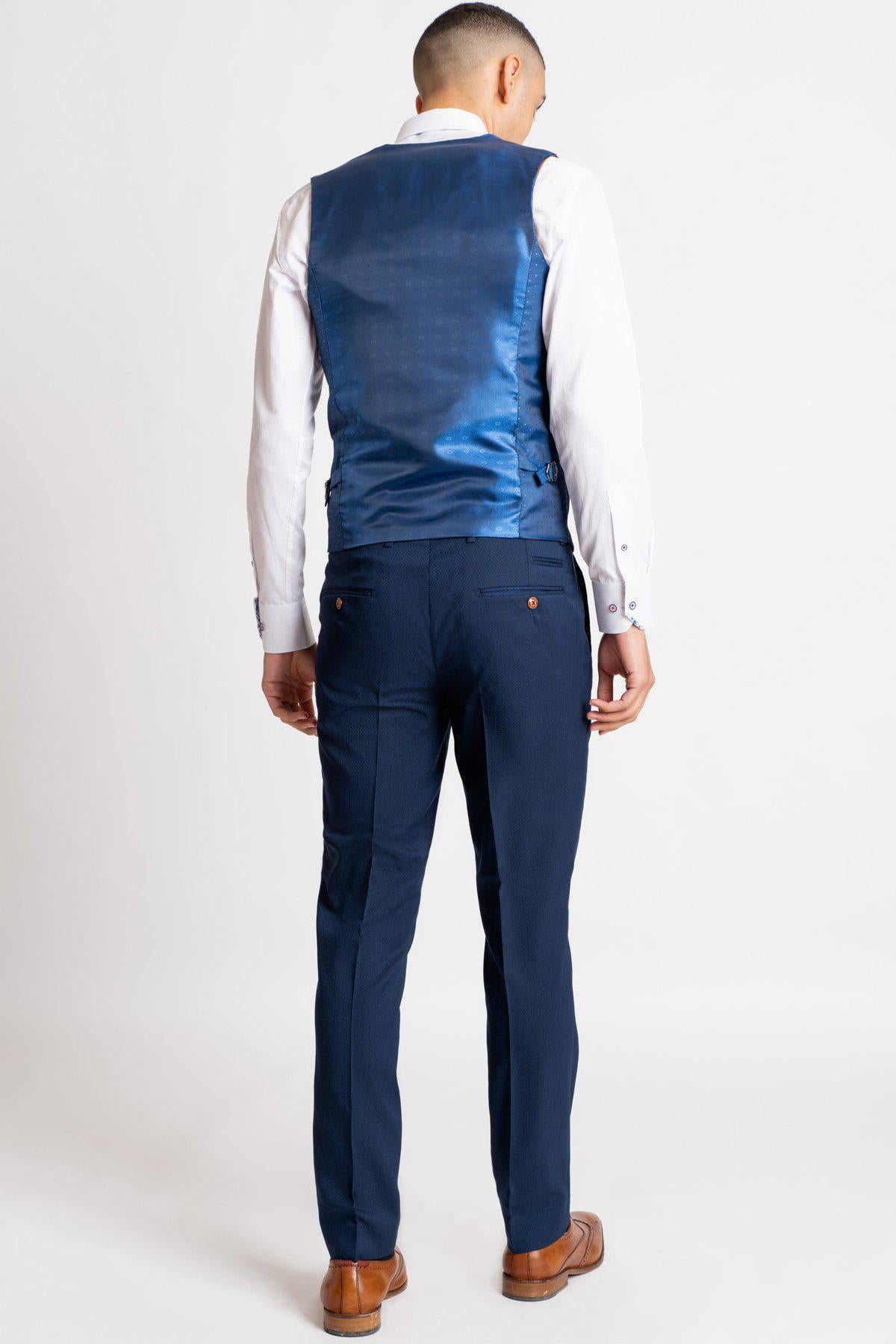 A man showcases The Everton Collection | James Garner In The MAX Royal Blue Suit by marcdarcy, pairing a royal blue vest over a white shirt, navy trousers, and brown shoes against a plain white background.