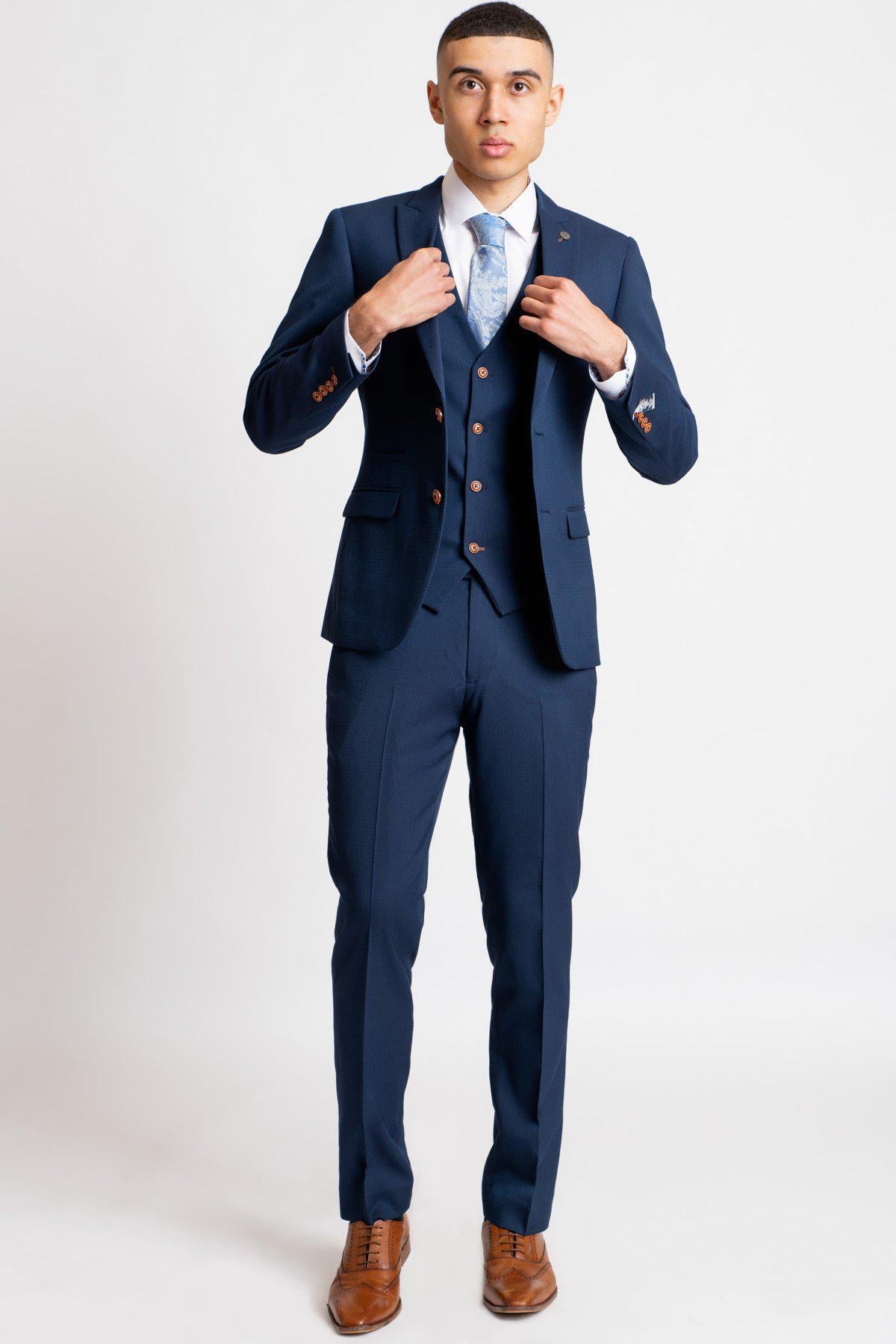 A young man models marcdarcy's The Everton Collection | James Garner in The MAX Royal Blue Suit, paired with a white shirt and light blue patterned tie, adjusting his jacket against a white background.