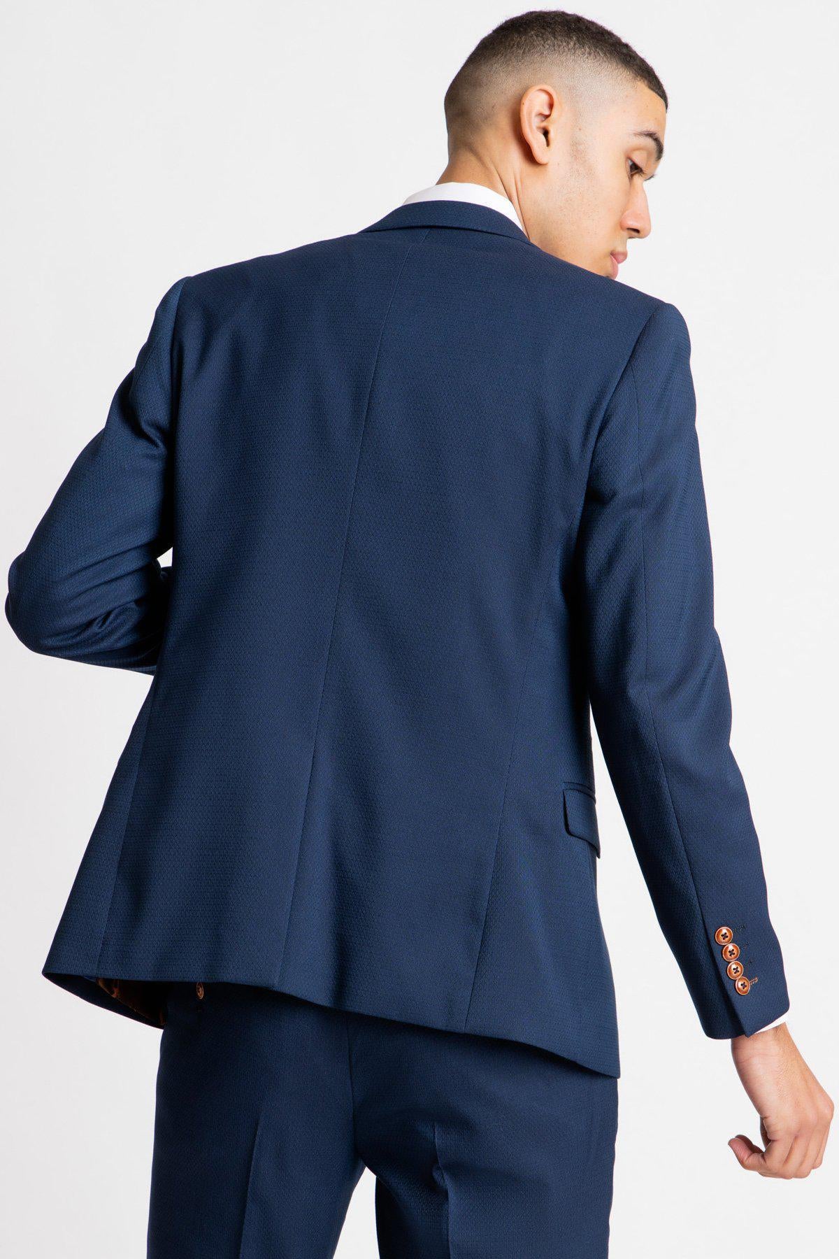 A person wears marcdarcy's The Everton Collection | James Garner In The MAX Royal Blue Suit, facing away from the camera and looking over their shoulder. The slim fit blazer has brown sleeve buttons against a white background.