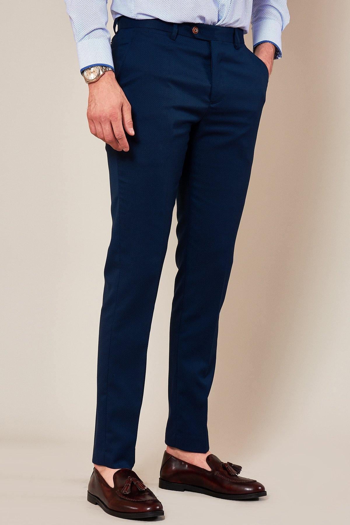 A man in marcdarcy MAX Skinny Fit Navy Trousers, a light blue dress shirt, a watch, and brown tassel loafers stands against a plain beige background with his hands relaxed at his sides.