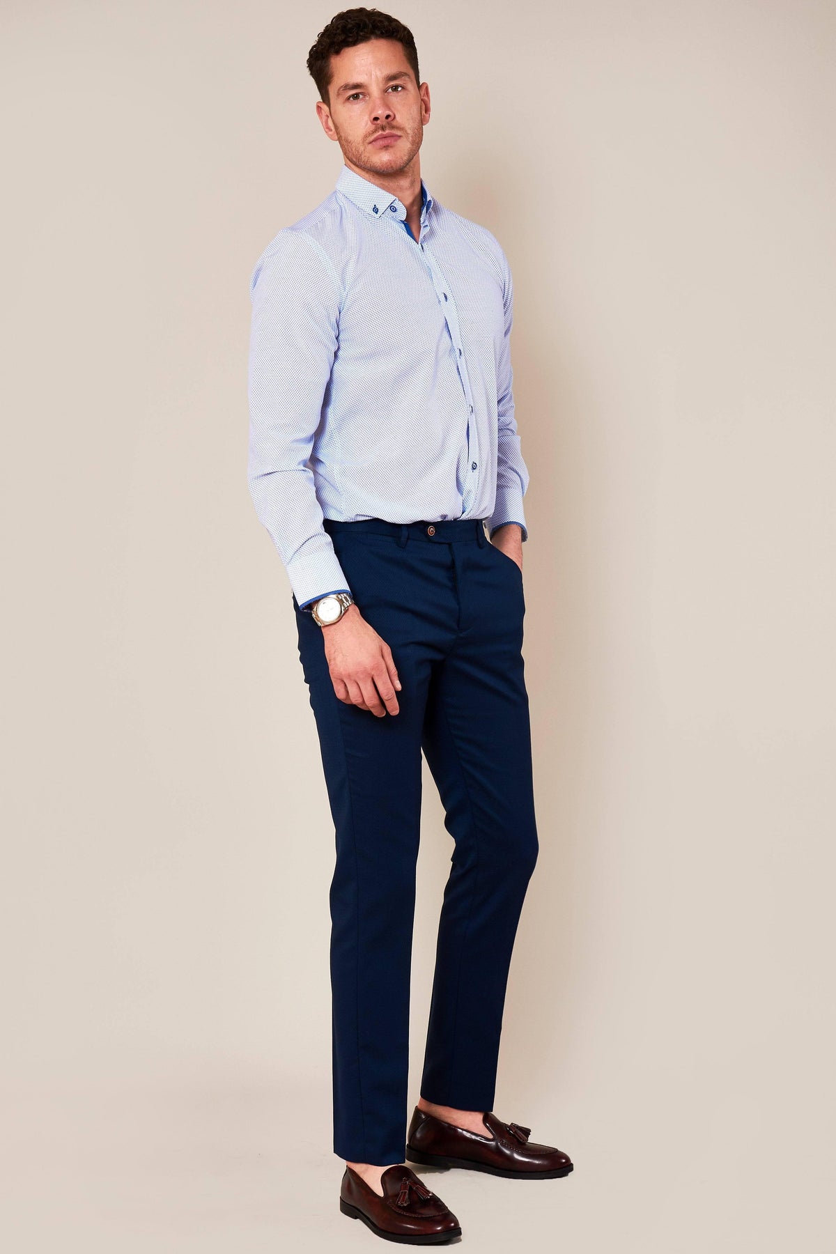 A man stands against a plain beige background wearing a light blue button-up shirt, marcdarcy MAX Skinny Fit Royal Blue Trousers, burgundy loafers, and a watch, with one hand in his pocket and a neutral expression.