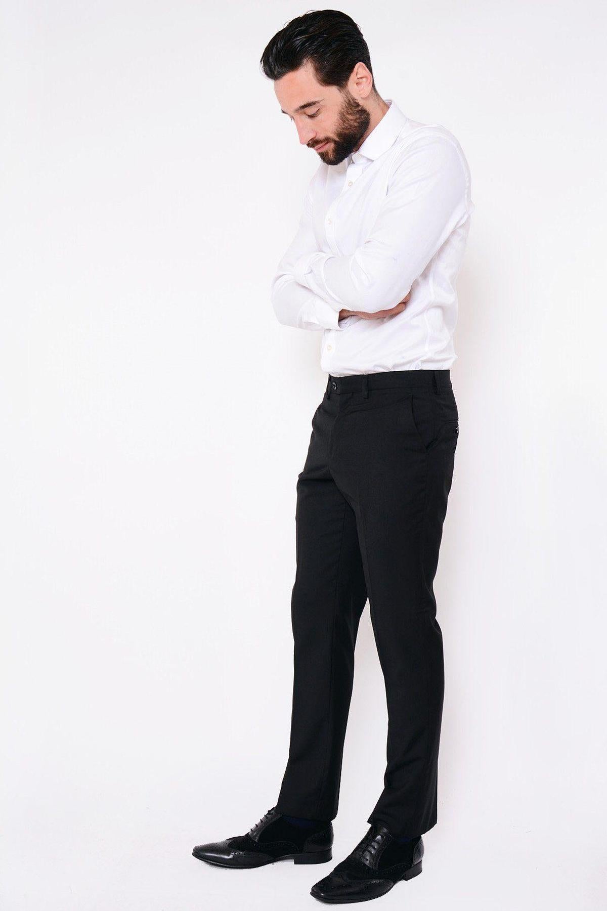 A bearded man stands against a white background, arms crossed and looking down, wearing a white shirt, black dress shoes, and marcdarcy RAMBO Black Flat Fronted Trousers.