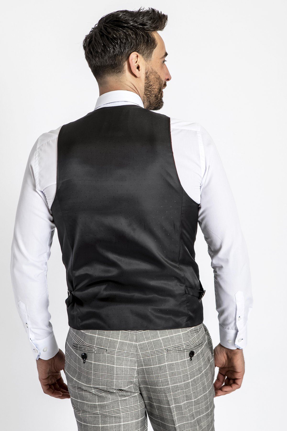 A man with short dark hair and a beard is seen from behind wearing a white shirt, grey plaid dress pants, and the marcdarcy RAMBO - Black Formal Dress Waistcoat, standing against a plain white background.