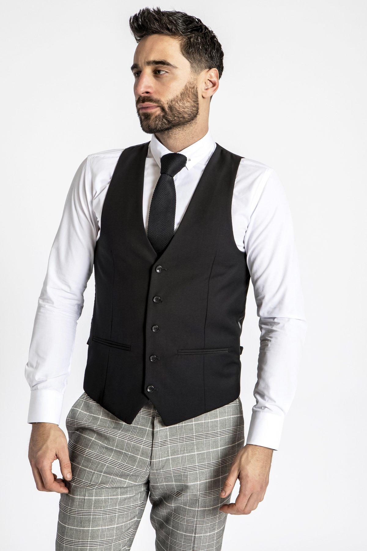 A man with short dark hair and a beard wears the marcdarcy RAMBO Black Formal Dress Waistcoat over a white shirt, paired with a black tie and gray plaid pants. He stands against a plain white background, looking off to the side.