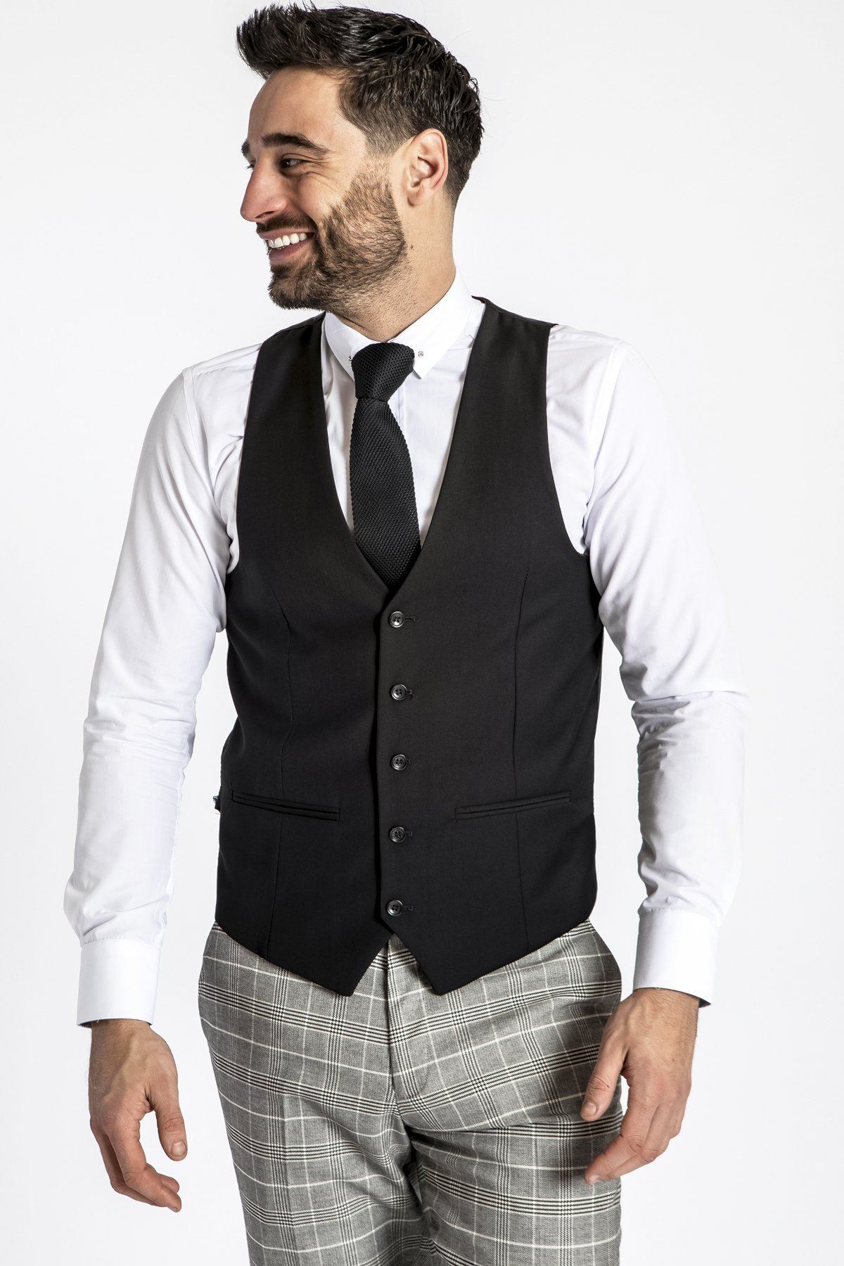 A man with short dark hair and a beard smiles, wearing a white dress shirt, black tie, gray plaid trousers, and the marcdarcy RAMBO Black Formal Dress Waistcoat against a plain white background.