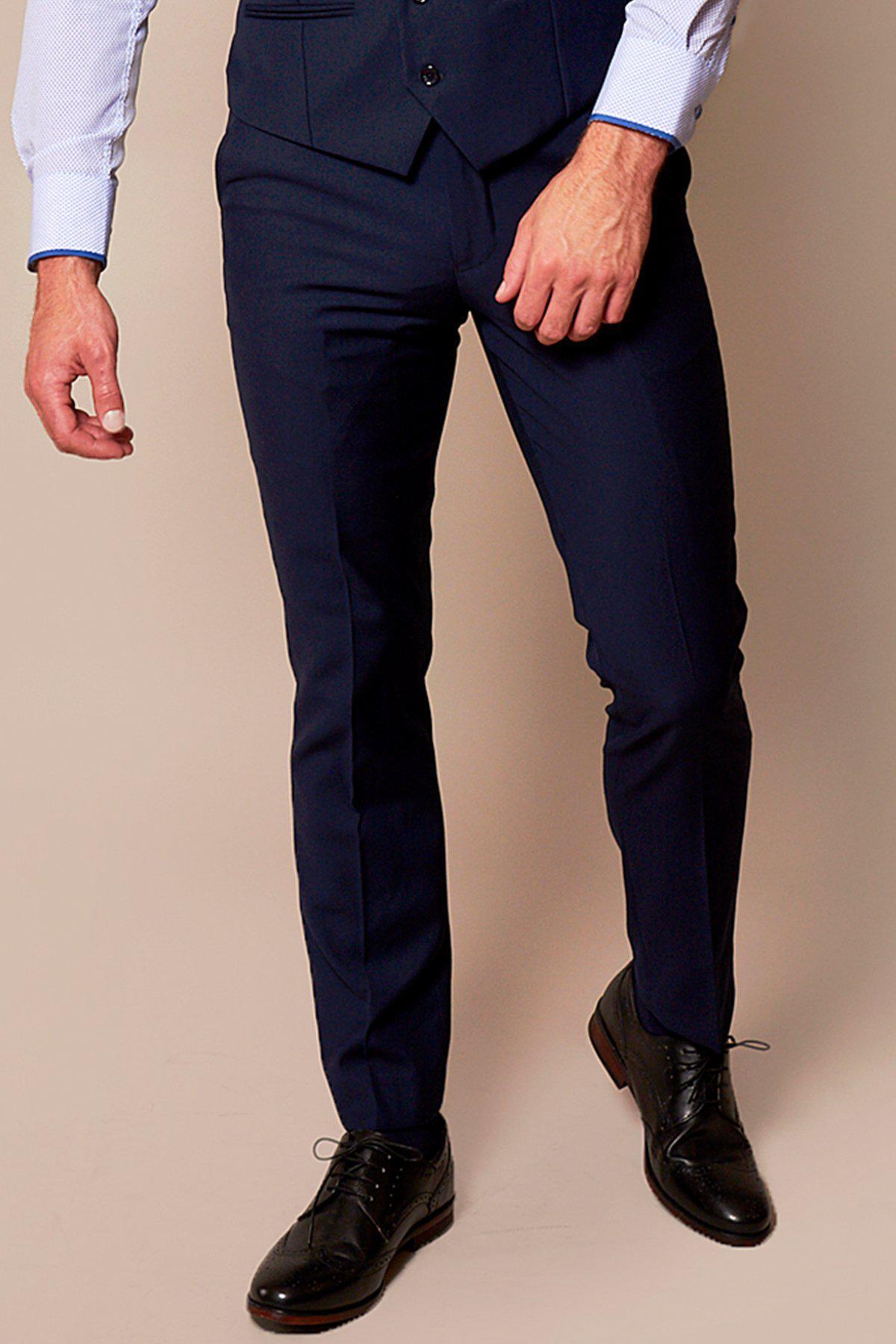 A man stands against a beige background in a navy suit, marcdarcy RAMBO navy flat fronted trousers, a light blue dress shirt, and black dress shoes. His pose highlights classic formalwear with his left hand by his side and right near his hip.