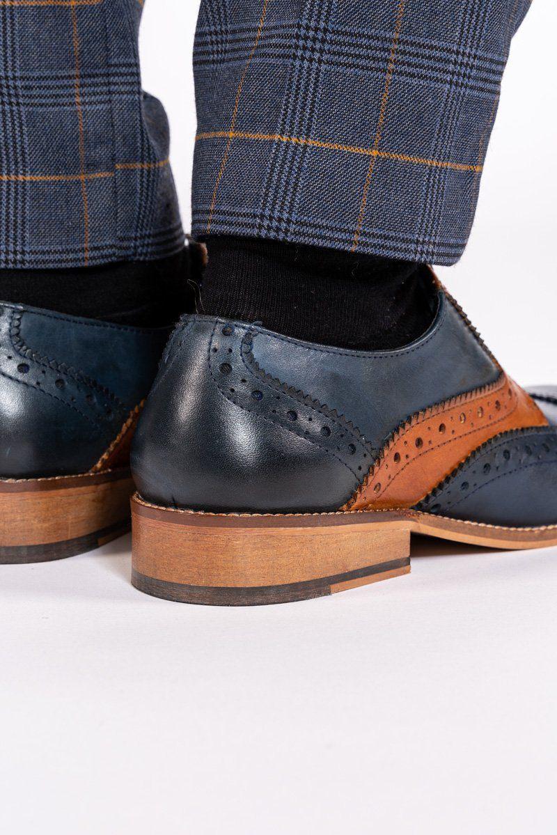 Close-up of a person wearing Marc Darcy RILEY Navy Tan Leather Contrast Oxford Brogue Shoes with black socks and blue plaid trousers with yellow lines, standing on a white surface.