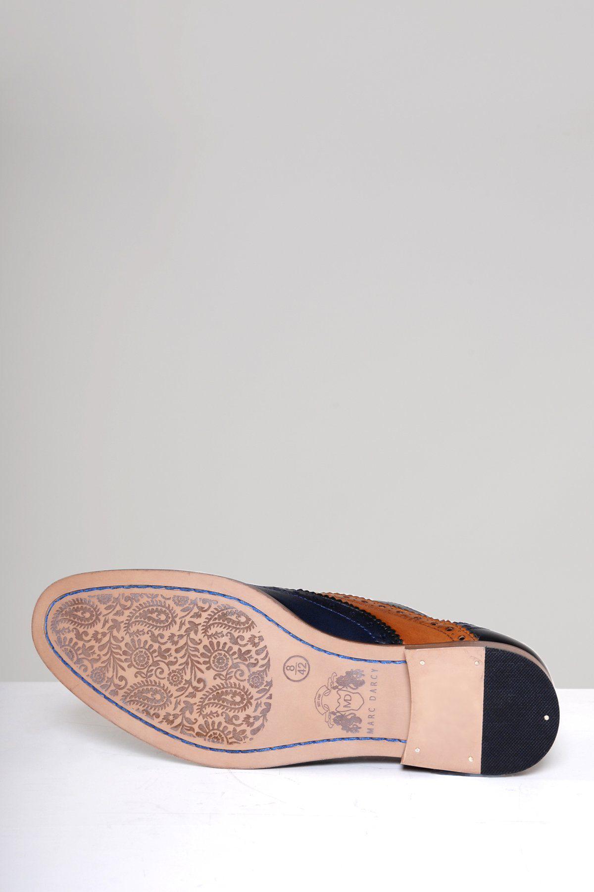 The Marc Darcy RILEY Navy Tan Leather Contrast Oxford Brogue Shoe lies on its side, revealing intricate floral patterns etched into the tan leather sole and subtle metal details near the heel.