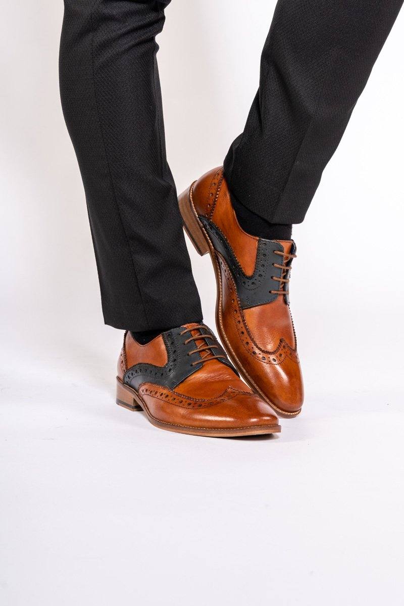 A person stands with one leg crossed over the other against a white background, wearing black pants and Marc Darcy’s RILEY Tan Navy Leather Contrast Oxford Brogue shoes.