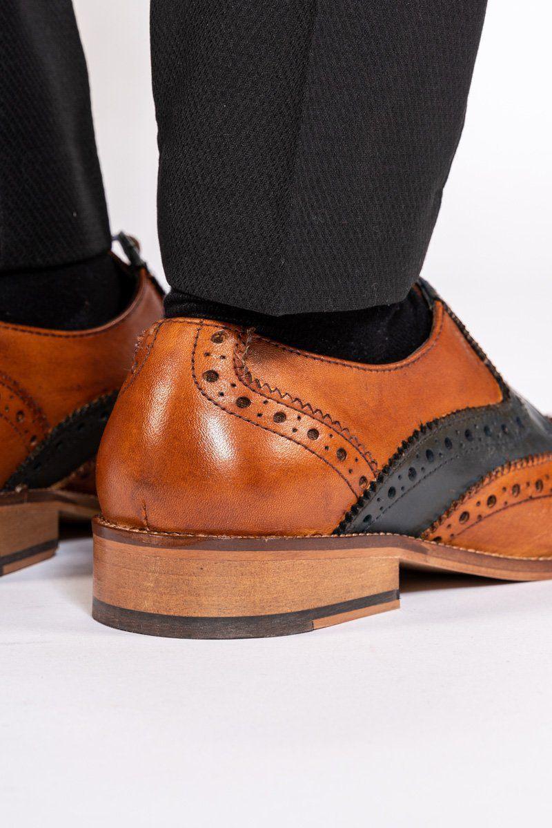 Close-up of a person wearing Marc Darcys RILEY Tan Navy Leather Contrast Oxford Brogue Shoes, featuring decorative perforations, wooden soles, and black trousers, standing on a white surface.