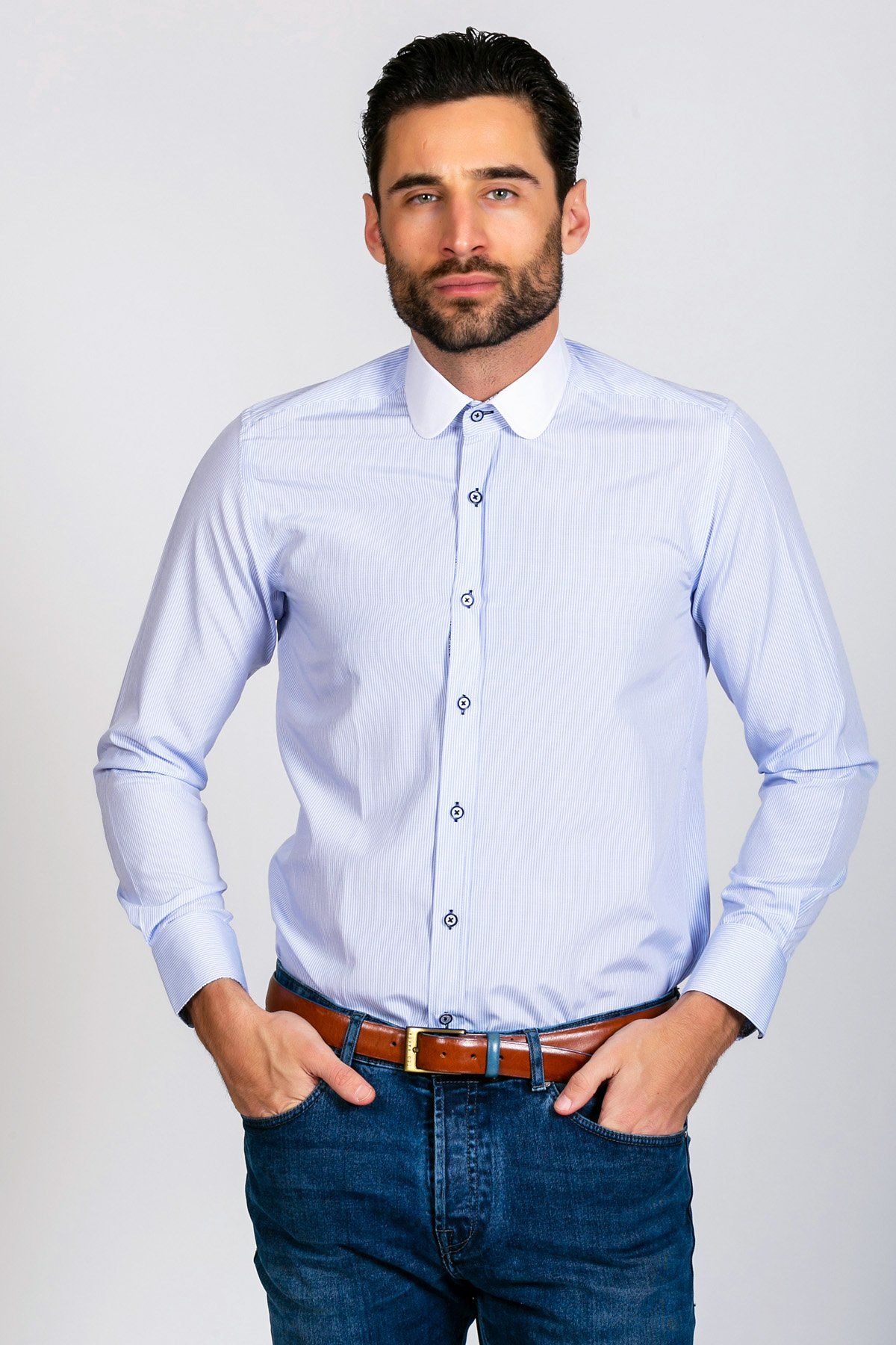 A man with dark hair and a beard wears the Marc Darcy SHELBY - Sky Blue Stripe Penny Collar Shirt, styled with blue jeans and a brown belt. He stands with hands in pockets against a plain light background.