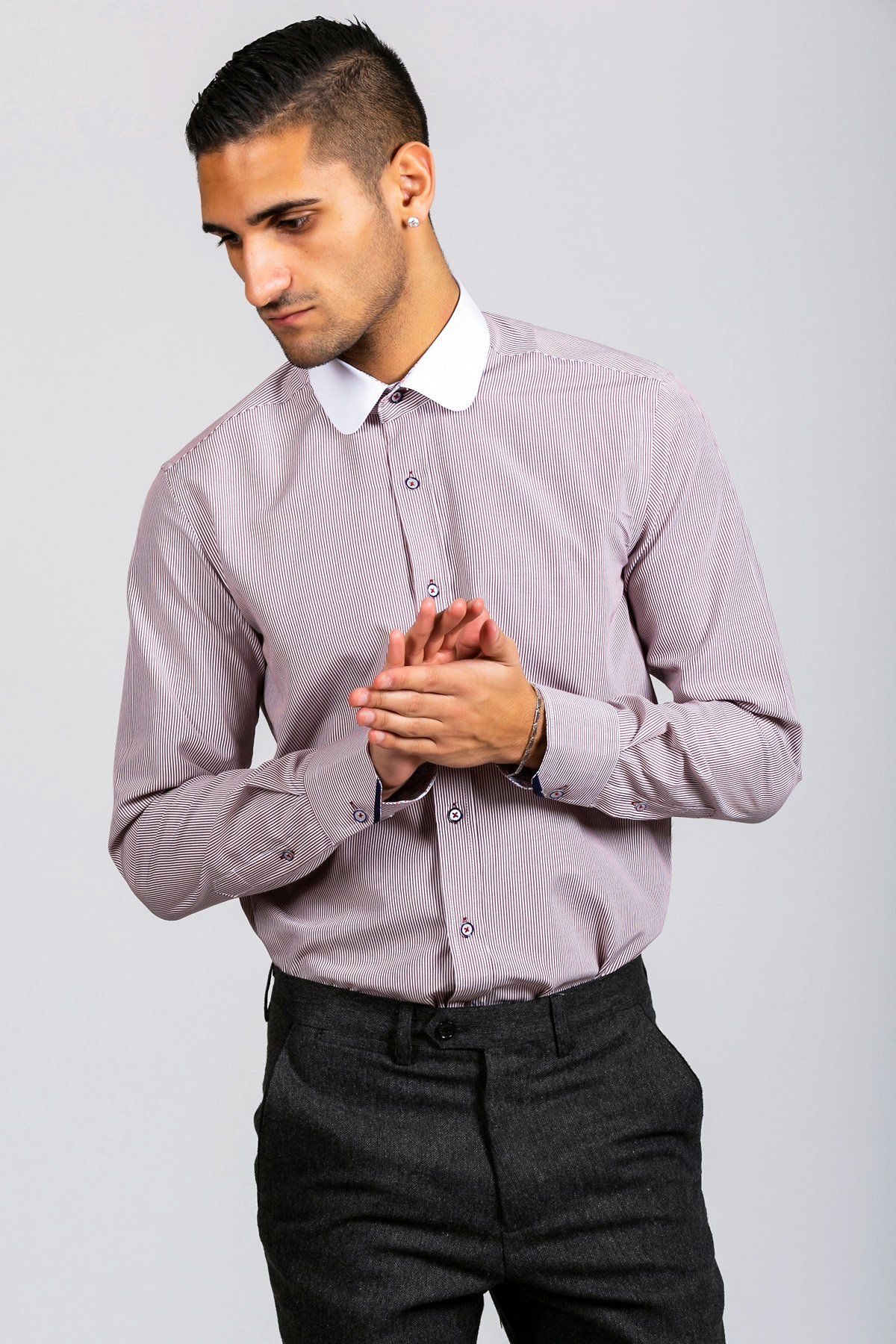 A style-conscious man with short dark hair wears the Marc Darcy SHELBY Wine Stripe Penny Collar Shirt and dark grey trousers, standing against a plain light background with his hands clasped together.
