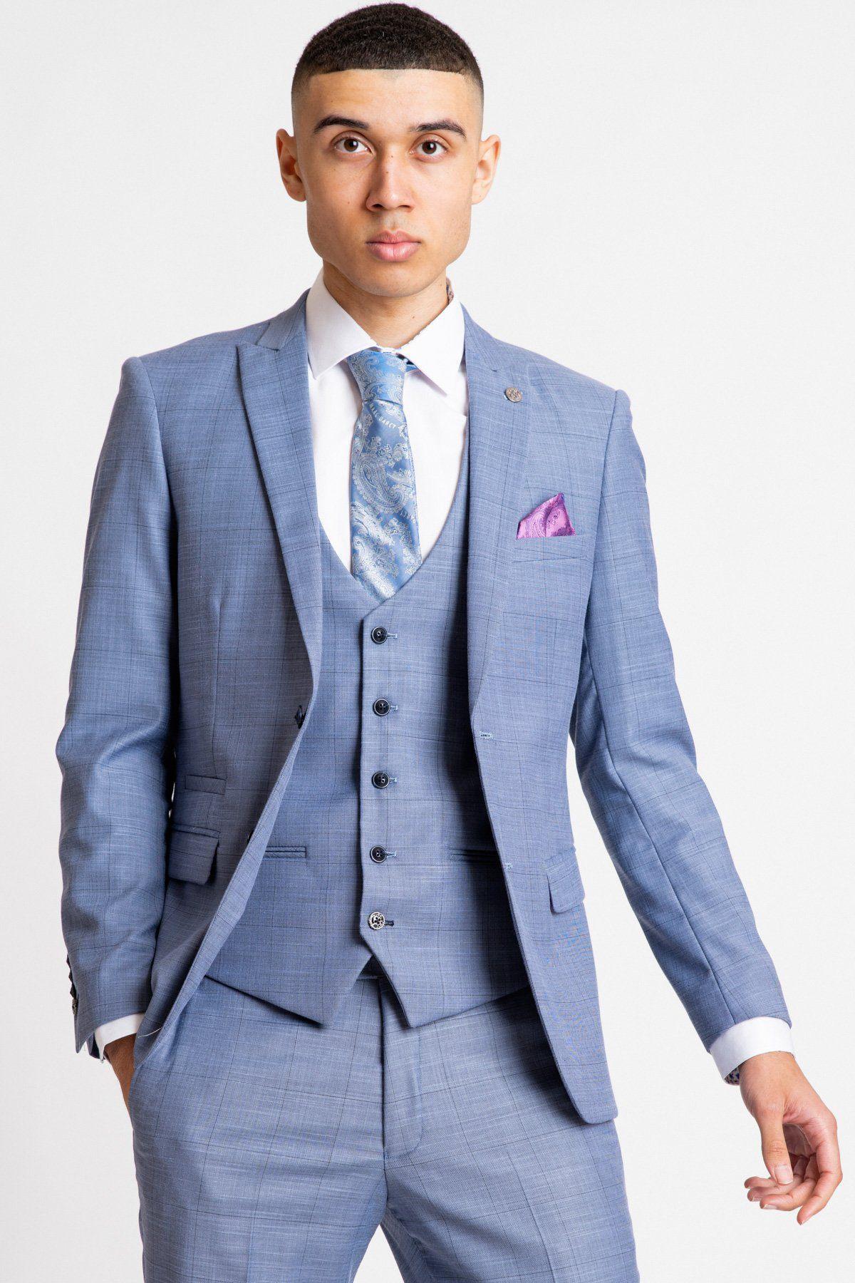 A young man wears the marcdarcy SID - Blue Grey Check Blazer as part of a three-piece suit with a white shirt, light blue patterned tie, and purple pocket square, standing with one hand in his pocket against a plain white background.