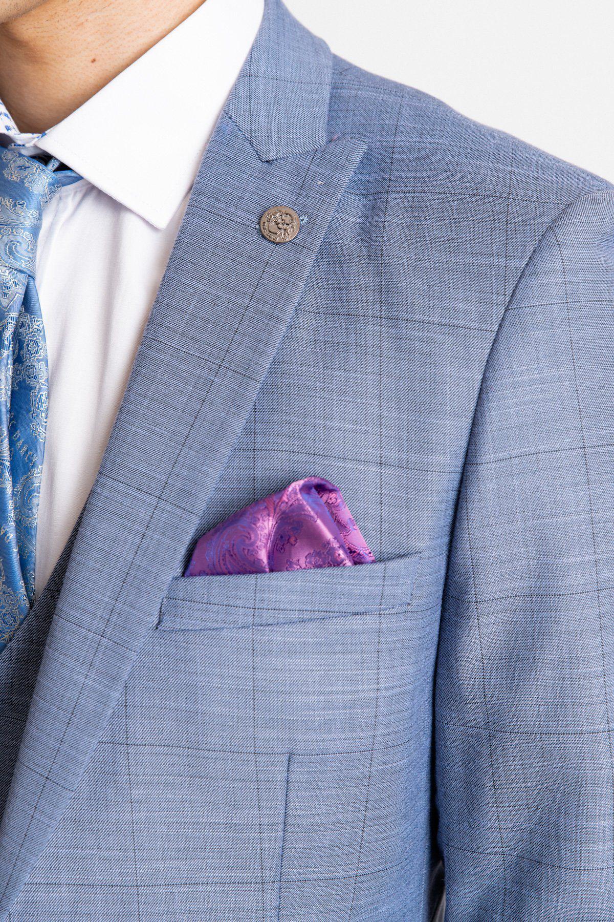 A close-up of a person wearing the marcdarcy SID - Blue Grey Check Blazer, styled with a white shirt, blue patterned tie, purple paisley pocket square, and decorative lapel pin.