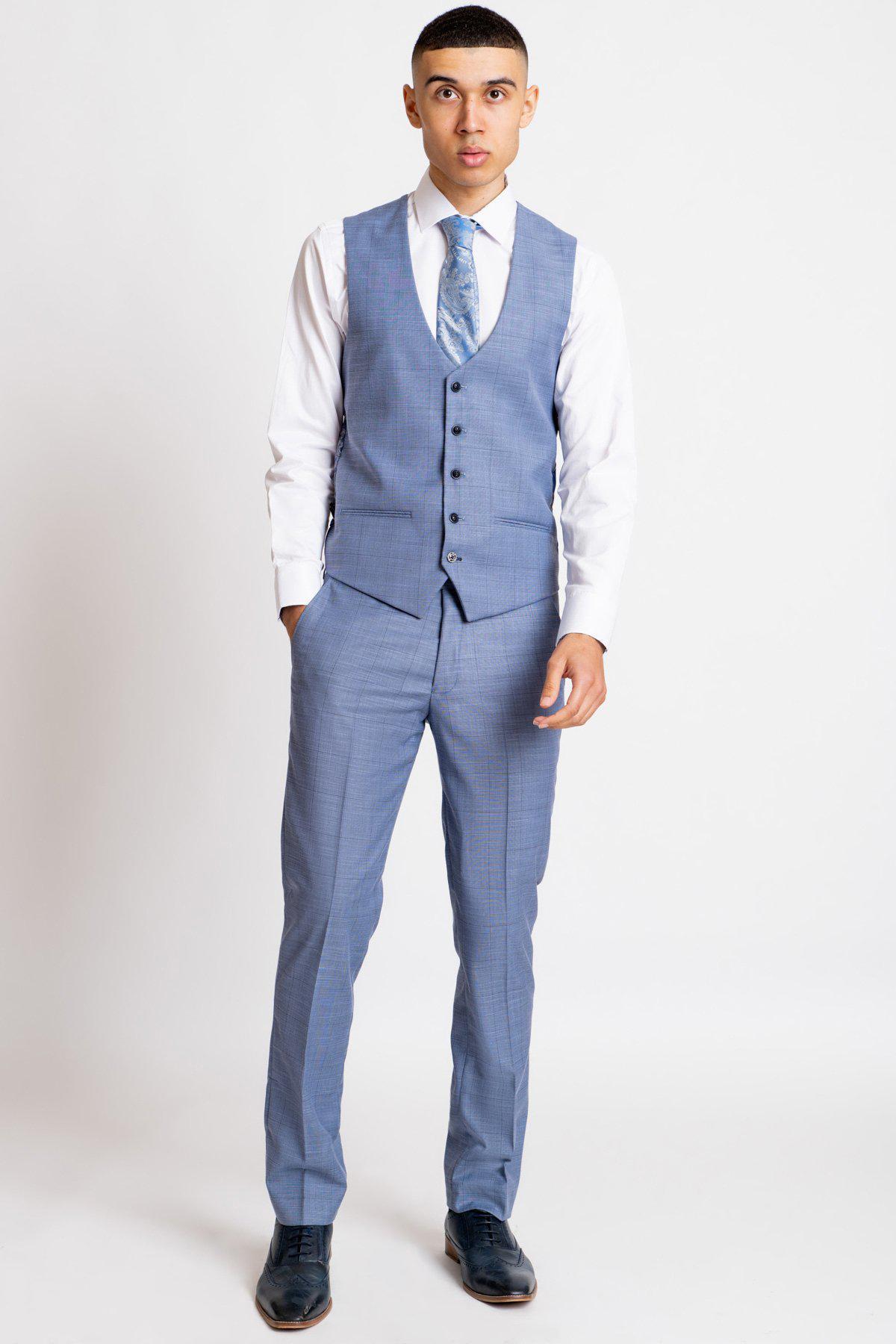 A young man stands against a plain white background, wearing the Marc Darcy SID Blue Grey Check Single Breasted Waistcoat as part of his three-piece suit, paired with a matching tie and white dress shirt. His left hand is in his pocket, looking at the camera.